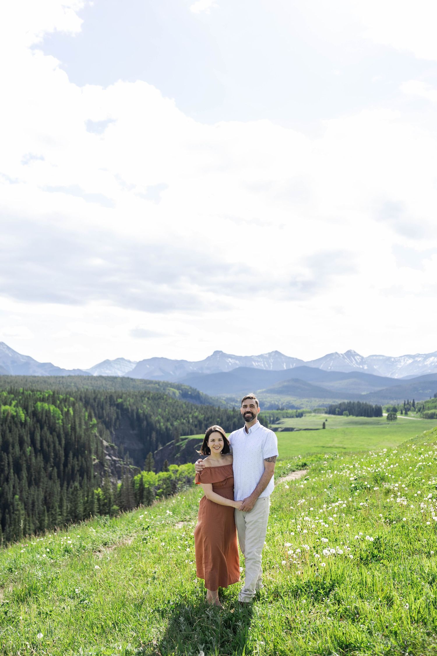 Couple embracing in a lush green mountain meadow with snow-capped peaks and evergreen forest backdrop.