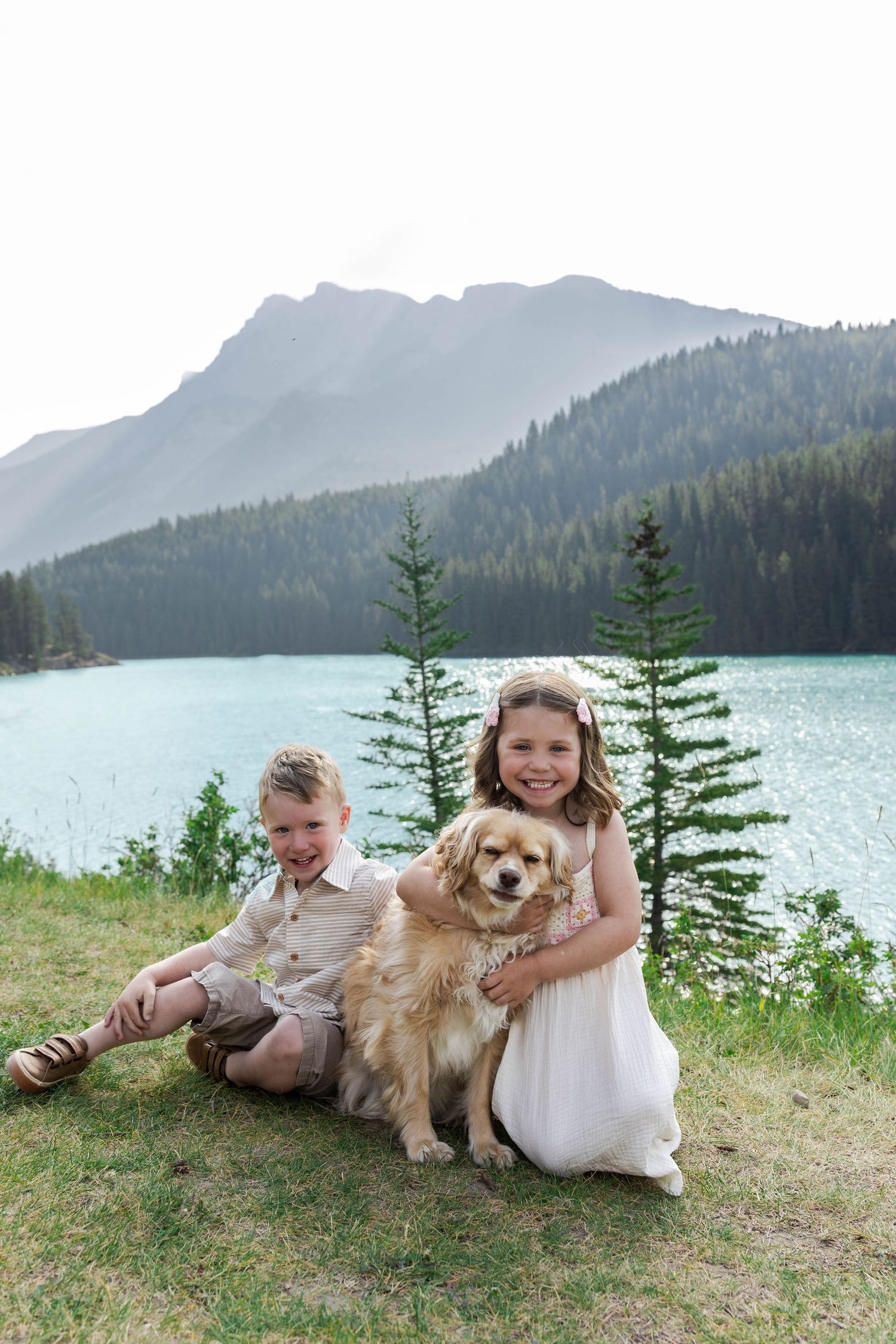 Two children and a golden dog sit on grass by a turquoise mountain Lake Minnewanka with pine trees in Banff