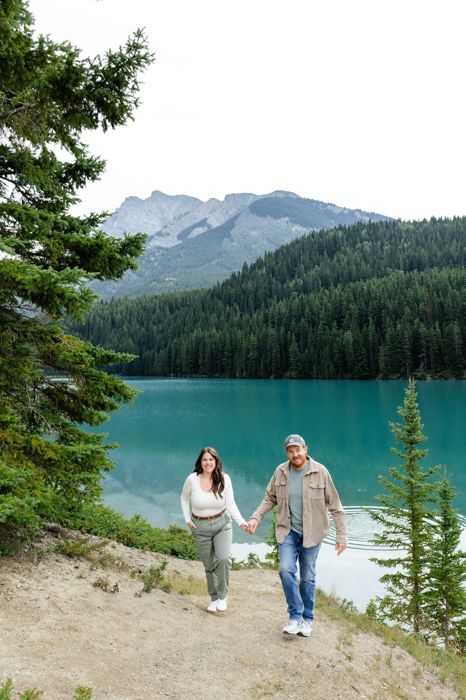 Couple holding hands walking along turquoise mountain lake trail surrounded by evergreen trees and rocky peaks.