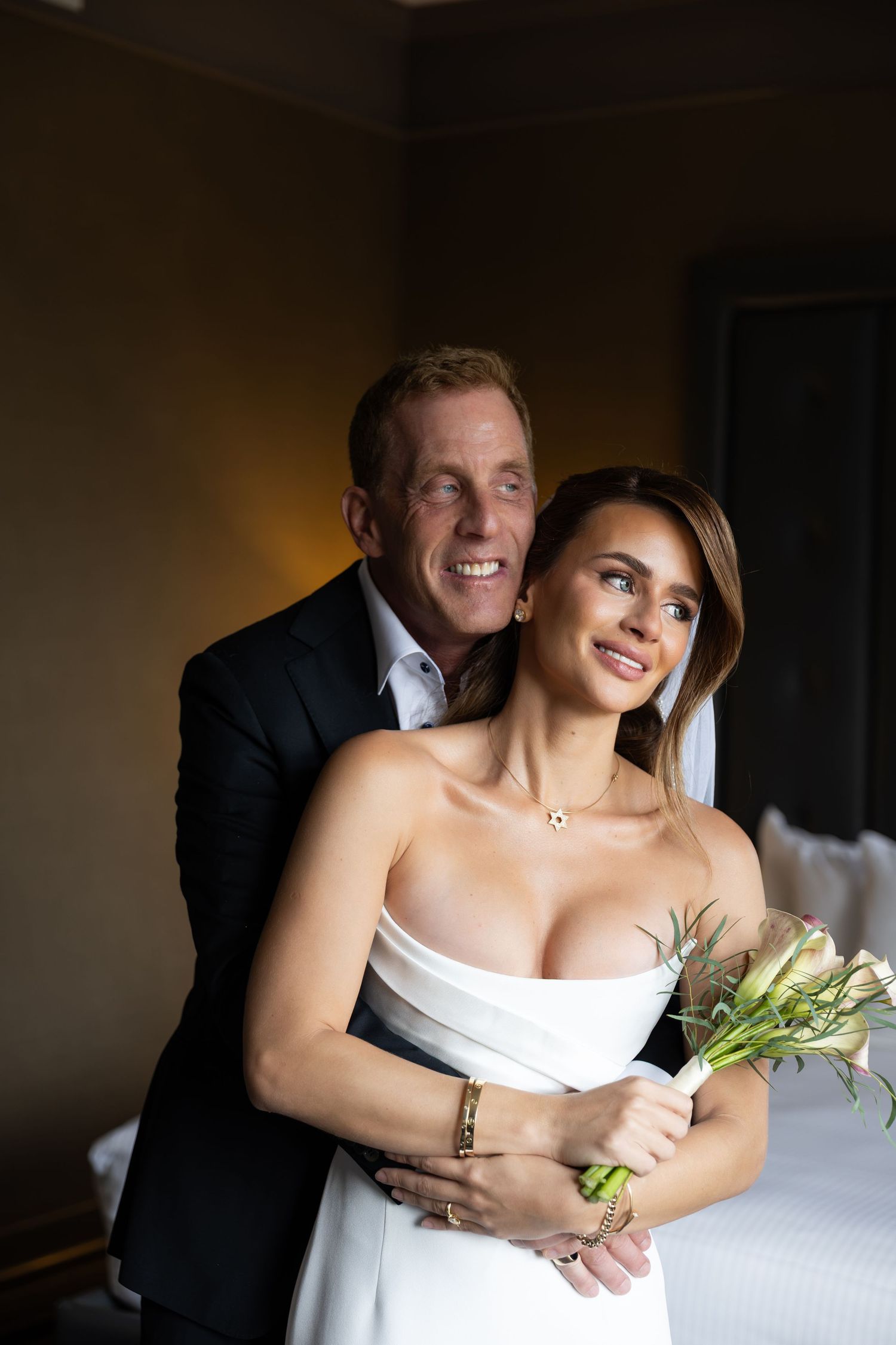 A smiling couple in wedding attire embraces indoors, the bride holding a bouquet in a white strapless gown.