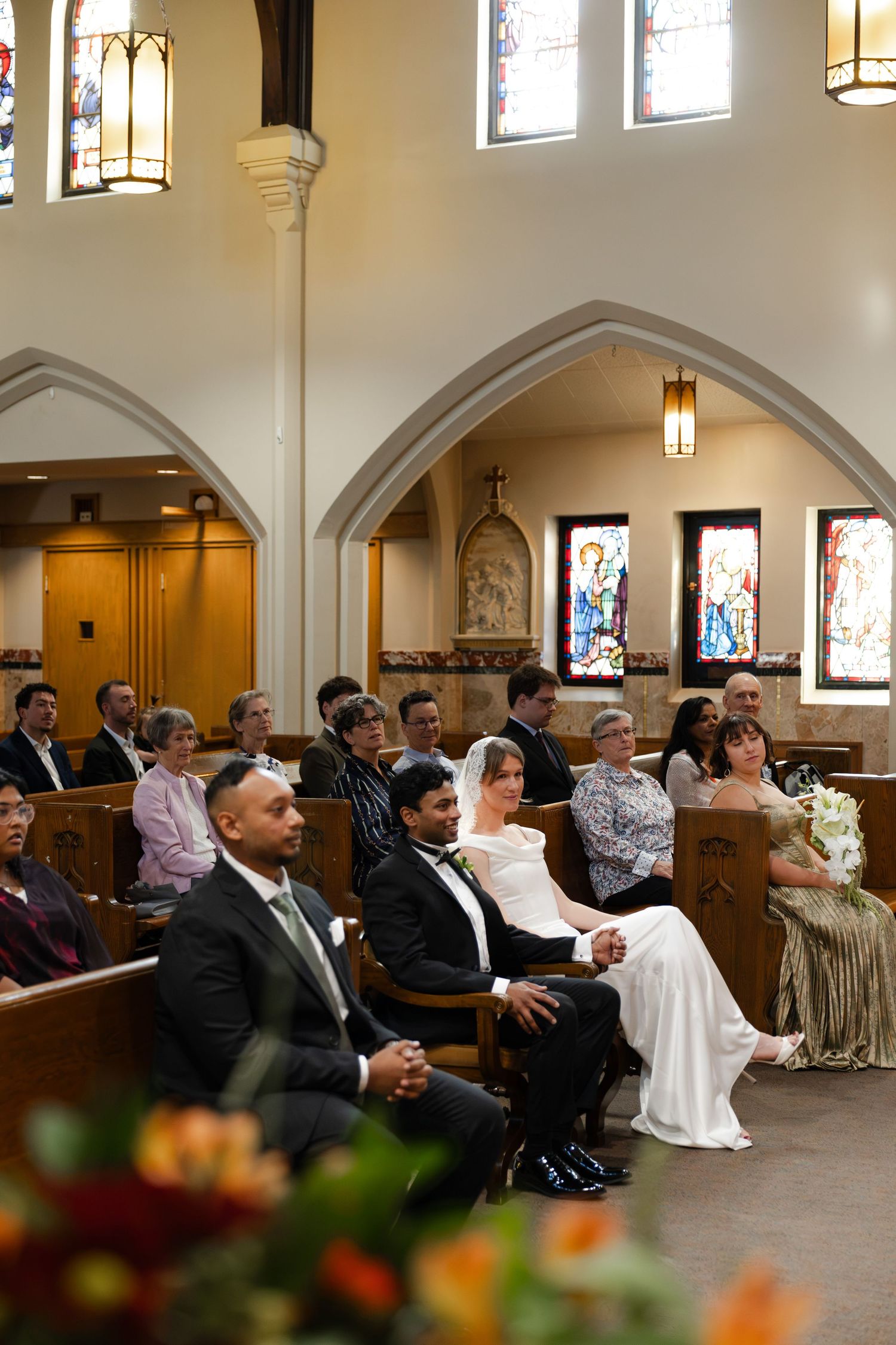 Bride and groom seated in church pews during wedding ceremony with guests and stained glass windows.