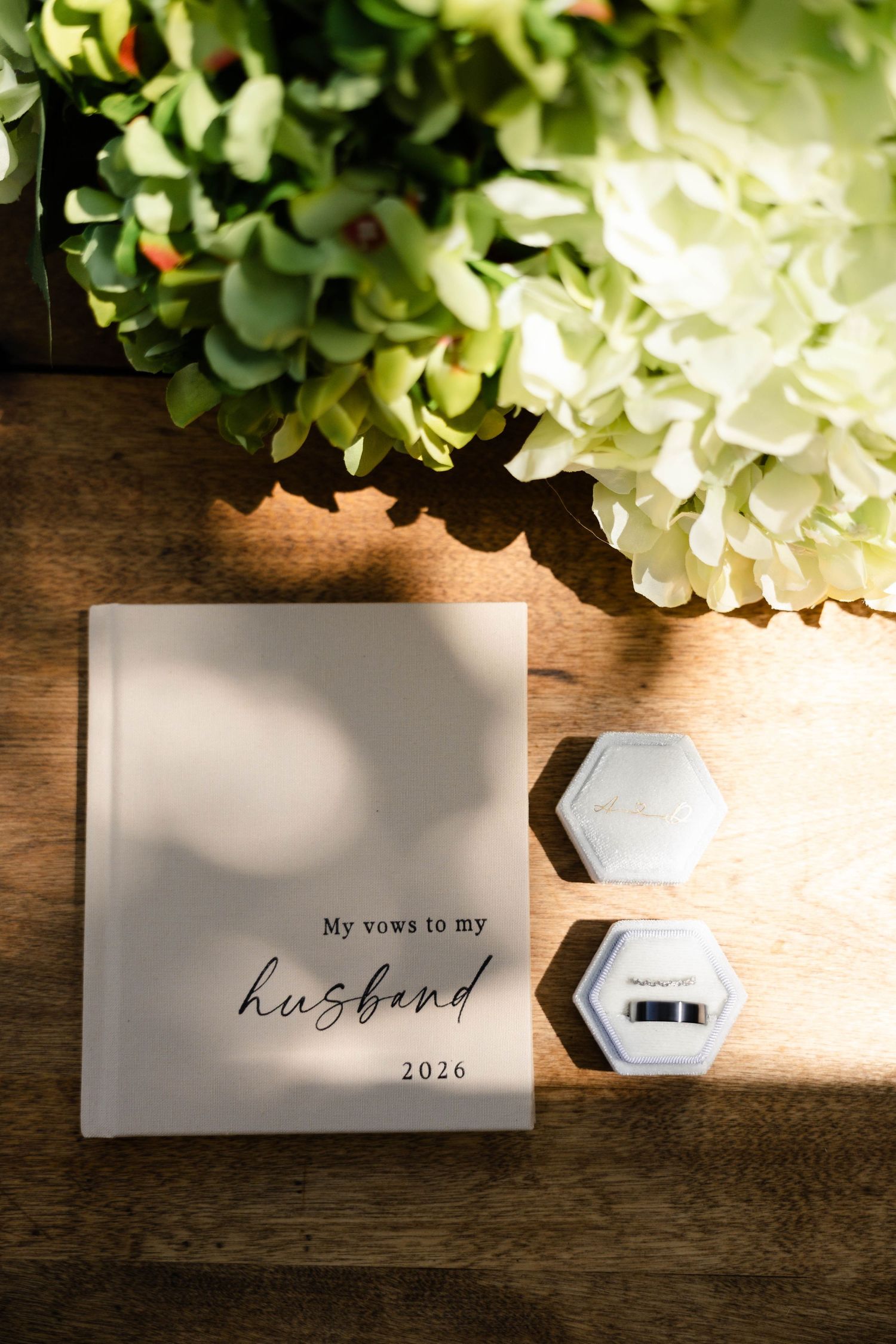 Wedding vow book reading 'My vows to my husband 2026' beside a hexagonal ring box and white hydrangeas on wood.