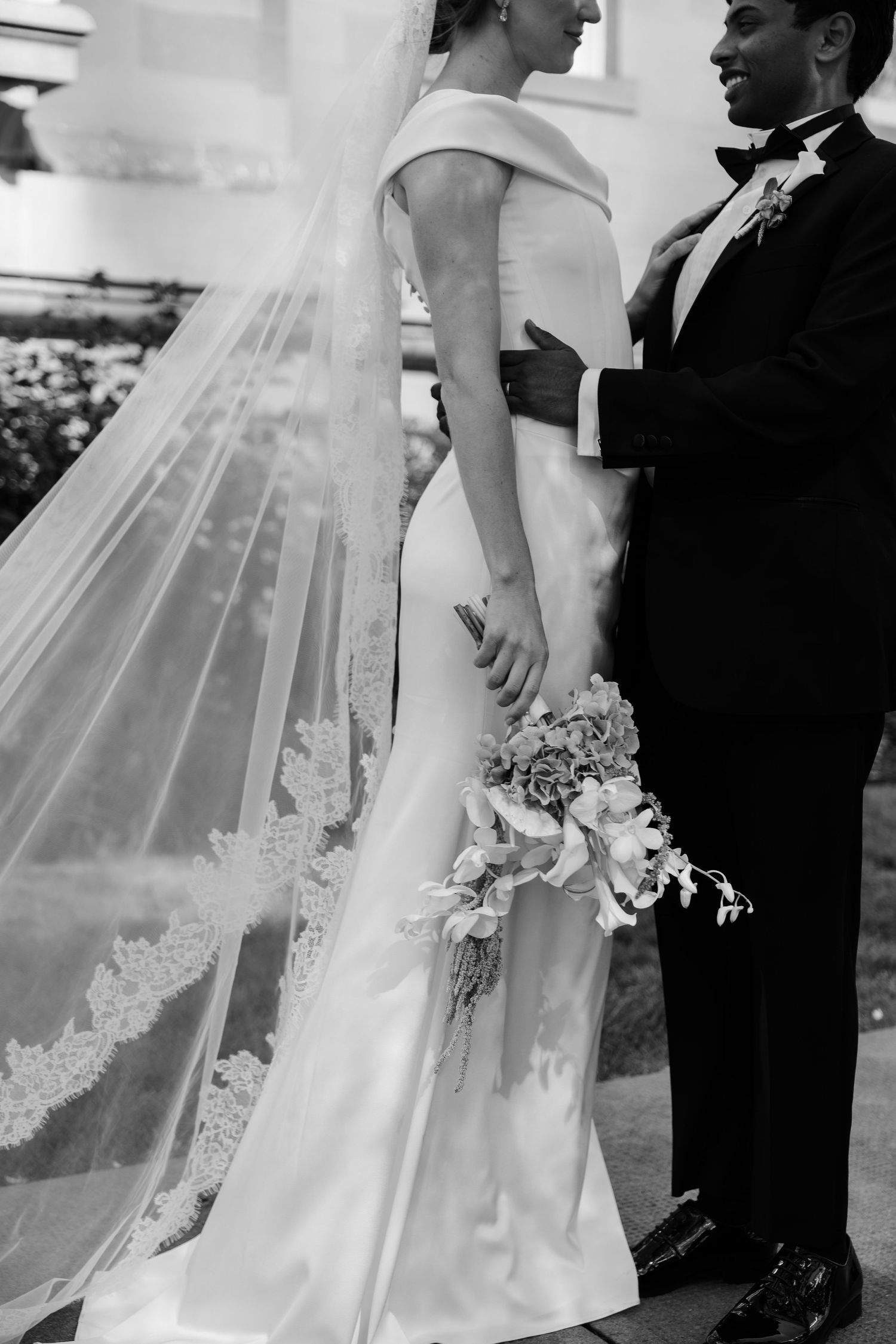 Black and white photo of bride in off-shoulder gown with lace veil holding bouquet with groom.