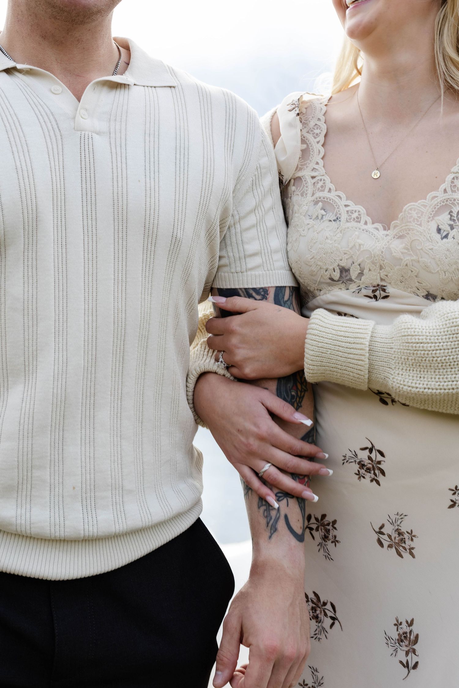 Couple embracing, man in cream knit polo and woman in lace floral dress, showing tattooed arm and engagement rings.Romantic couple in Minnew