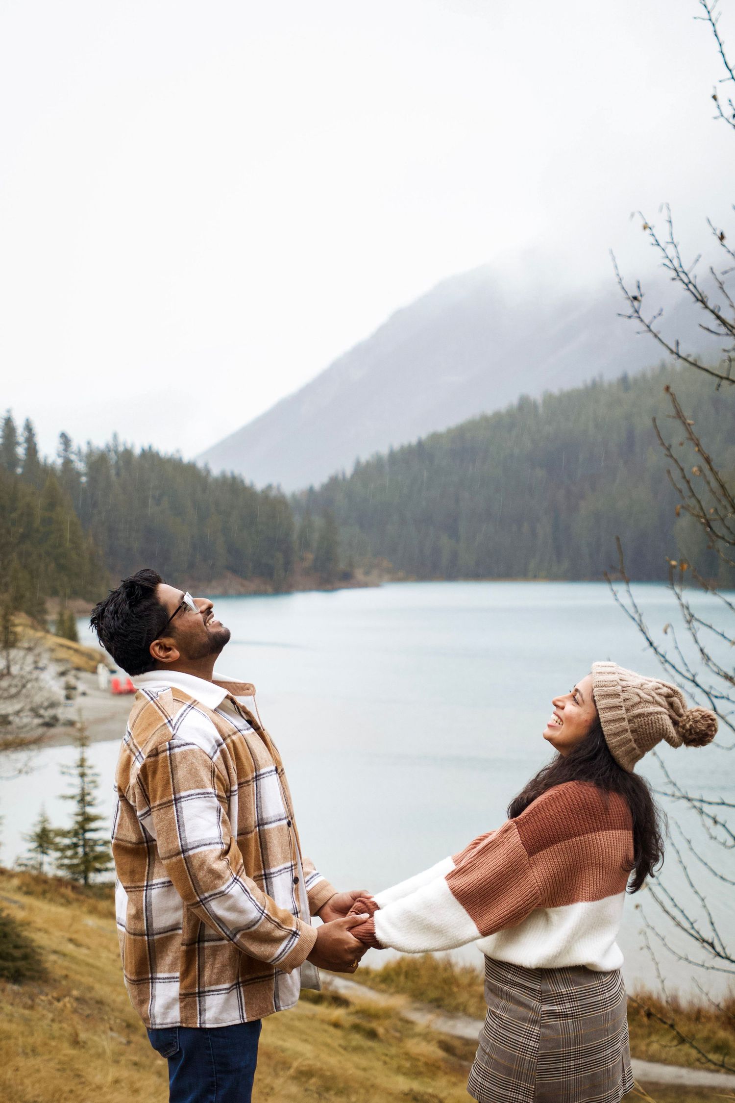 Couple holding hands by a misty mountain lake surrounded by evergreen forest on a cloudy day.