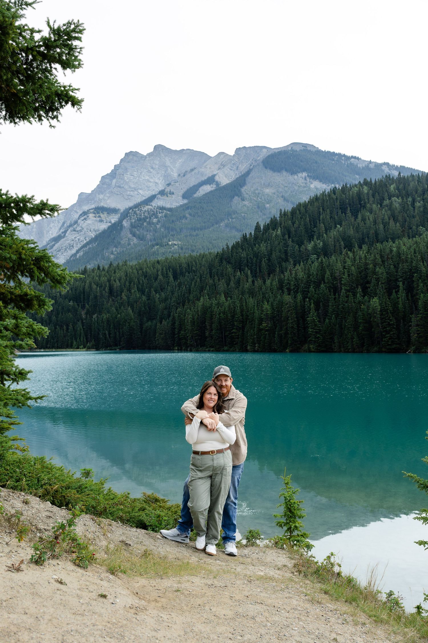 Couple embracing by turquoise mountain lake with evergreen forest and rocky peaks in Banff, Canada.