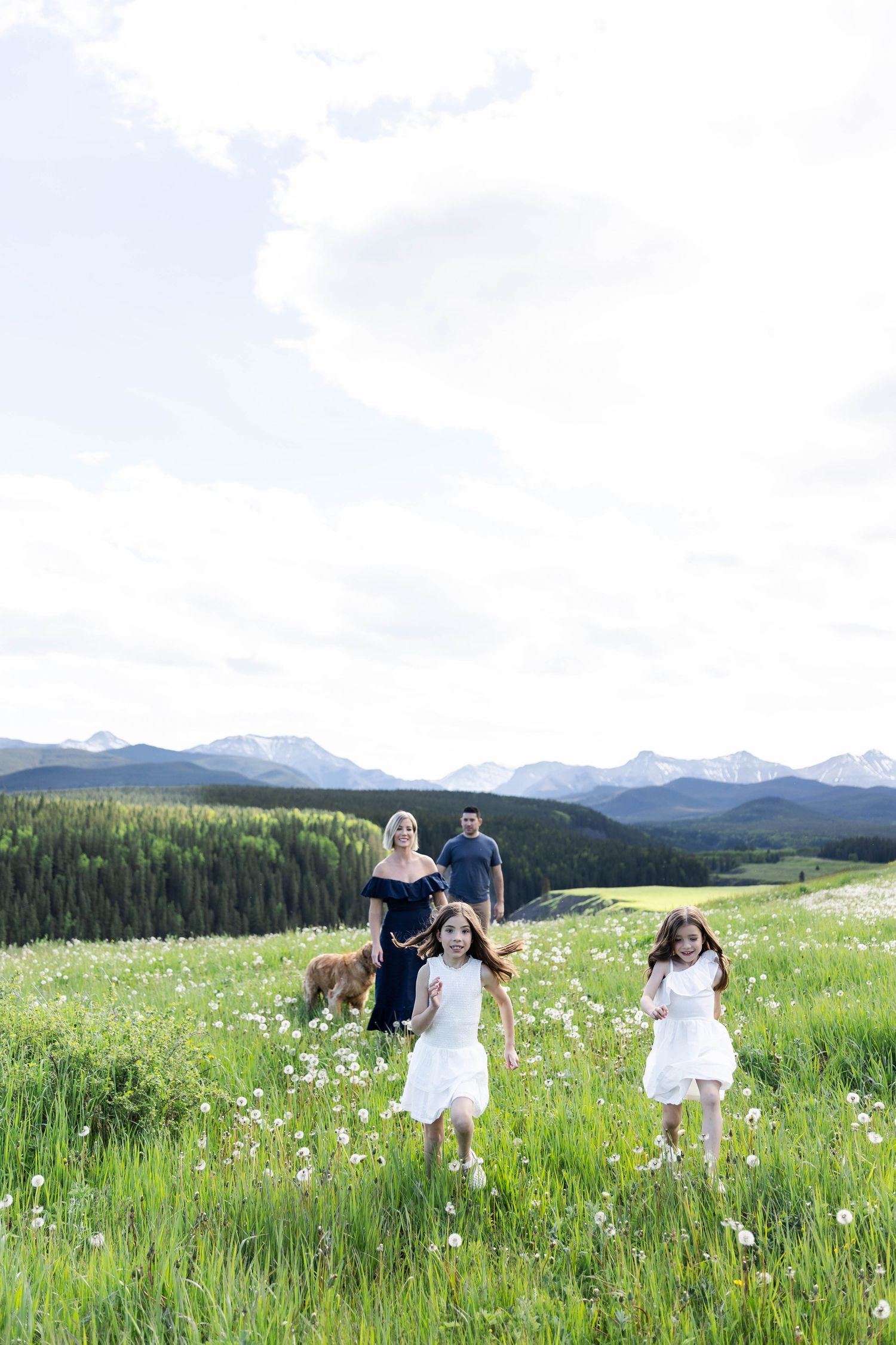 Family with two girls in white dresses running through a mountain meadow with wildflowers and snow-capped peaks.