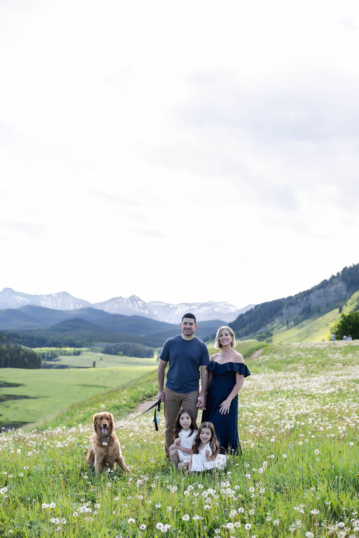 Family with two girls and golden retriever posing in wildflower meadow with snow-capped mountains backdrop.