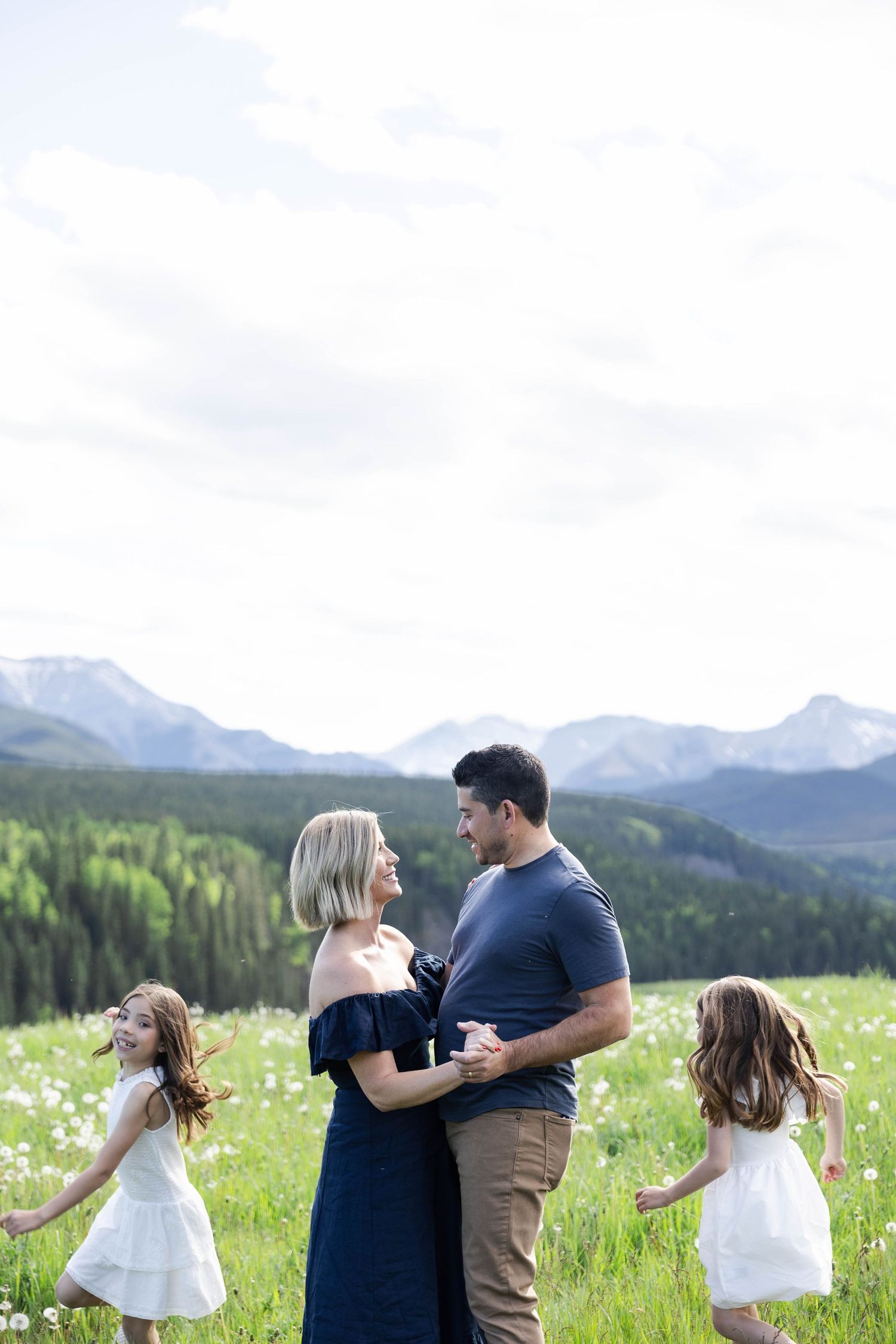 Family dancing in a wildflower meadow with snow-capped mountains, two girls in white dresses spinning around their parents.