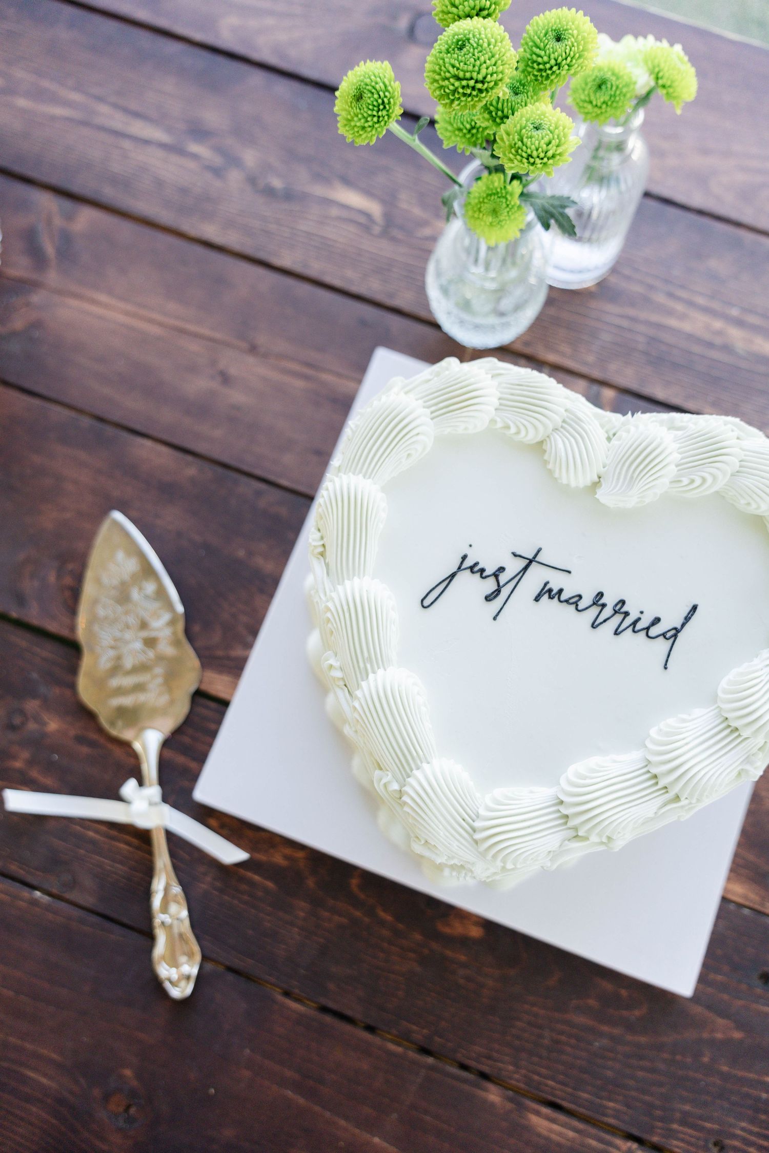 White heart-shaped wedding cake with just married text, cake server, and green flowers on rustic wood table.
