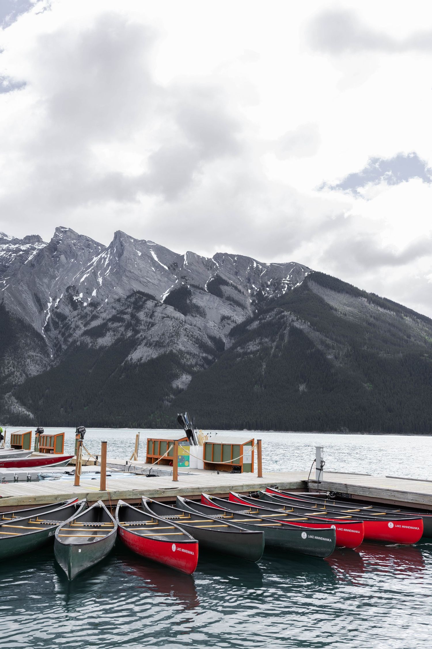 Red and green canoes docked on a mountain lake pier with snow-capped peaks and cloudy skies in the background.