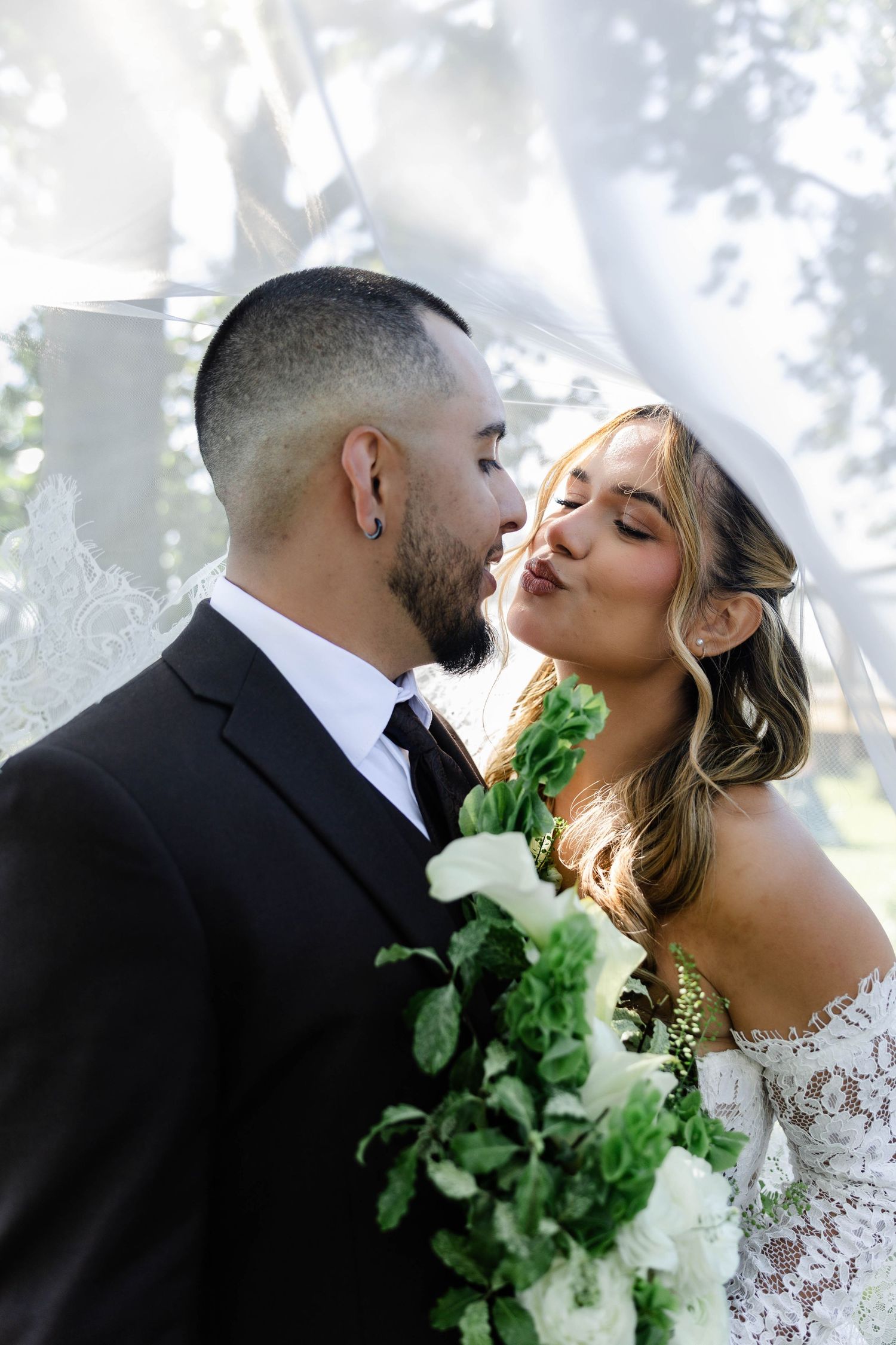 Bride and groom share a romantic kiss under a lace veil, bride holding a lush white and green bouquet.