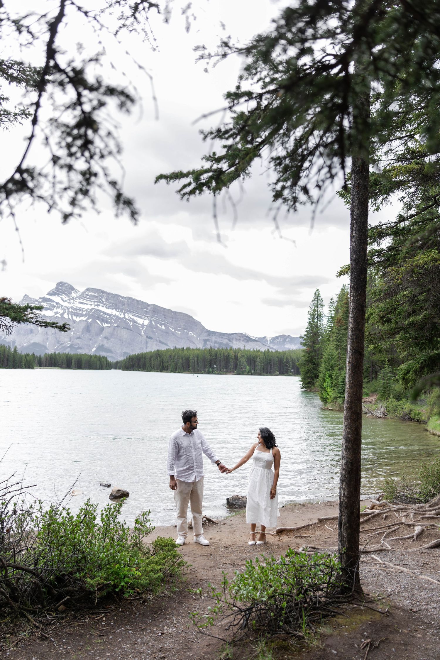 Engaged couple holding hands by a mountain lake in Banff, surrounded by pine trees and snow-capped peaks.