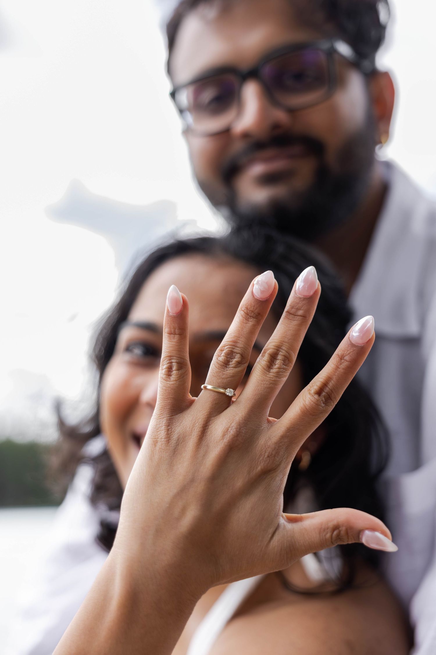 Couple joyfully displaying engagement ring with diamond solitaire, woman showing hand to camera with French manicure.