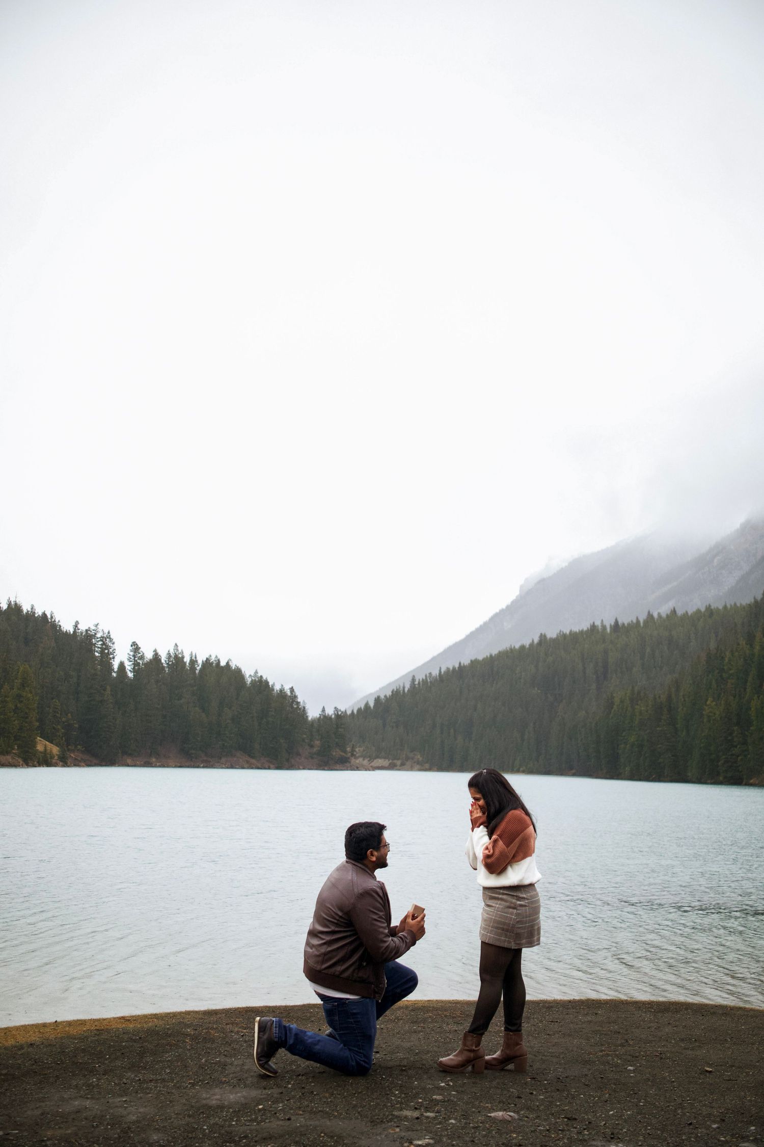 Man proposes on one knee to woman by a misty mountain lake surrounded by evergreen forests.