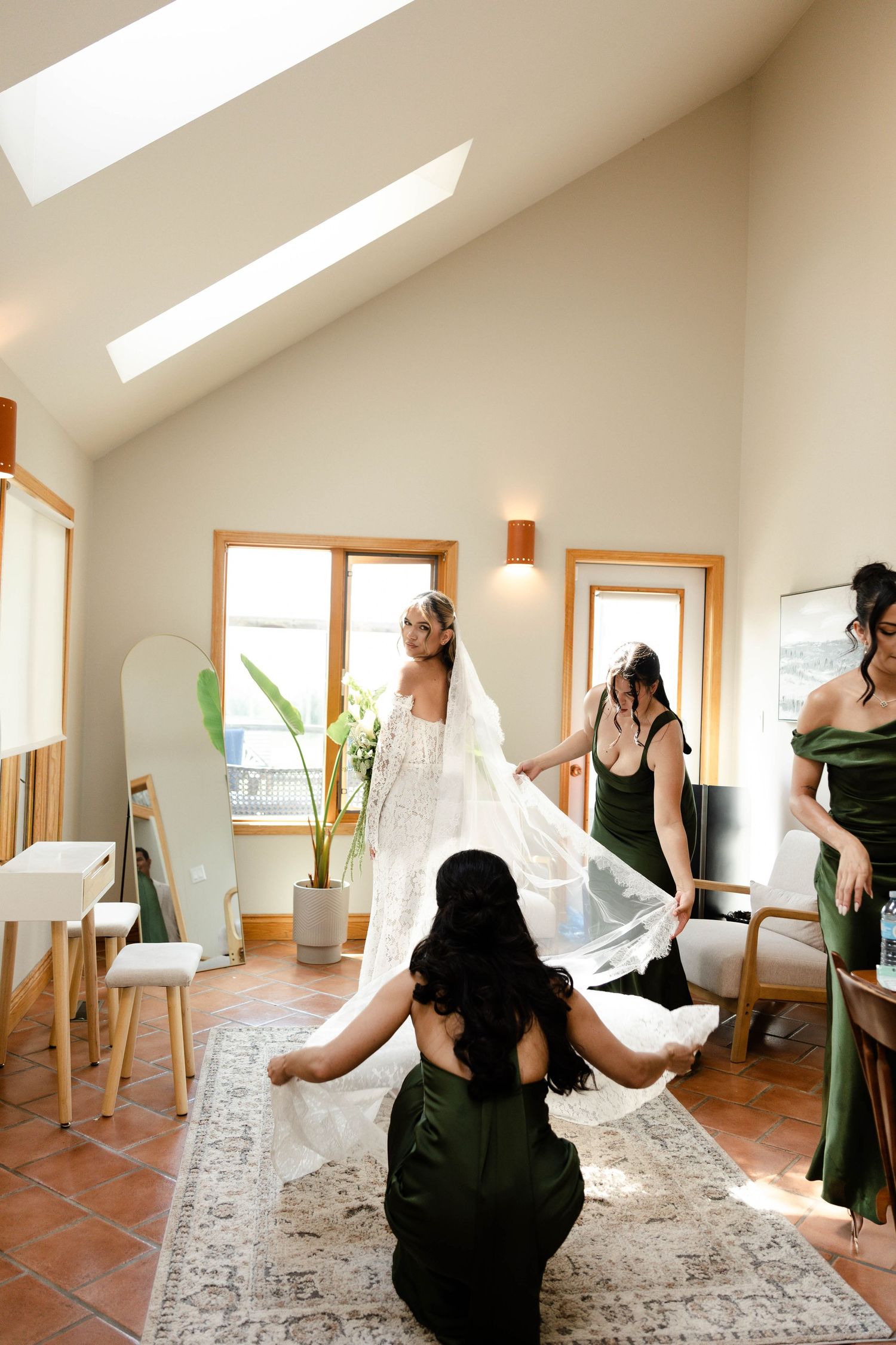 Bride in lace gown with bridesmaids in green dresses adjusting her veil in a bright, modern bridal suite.