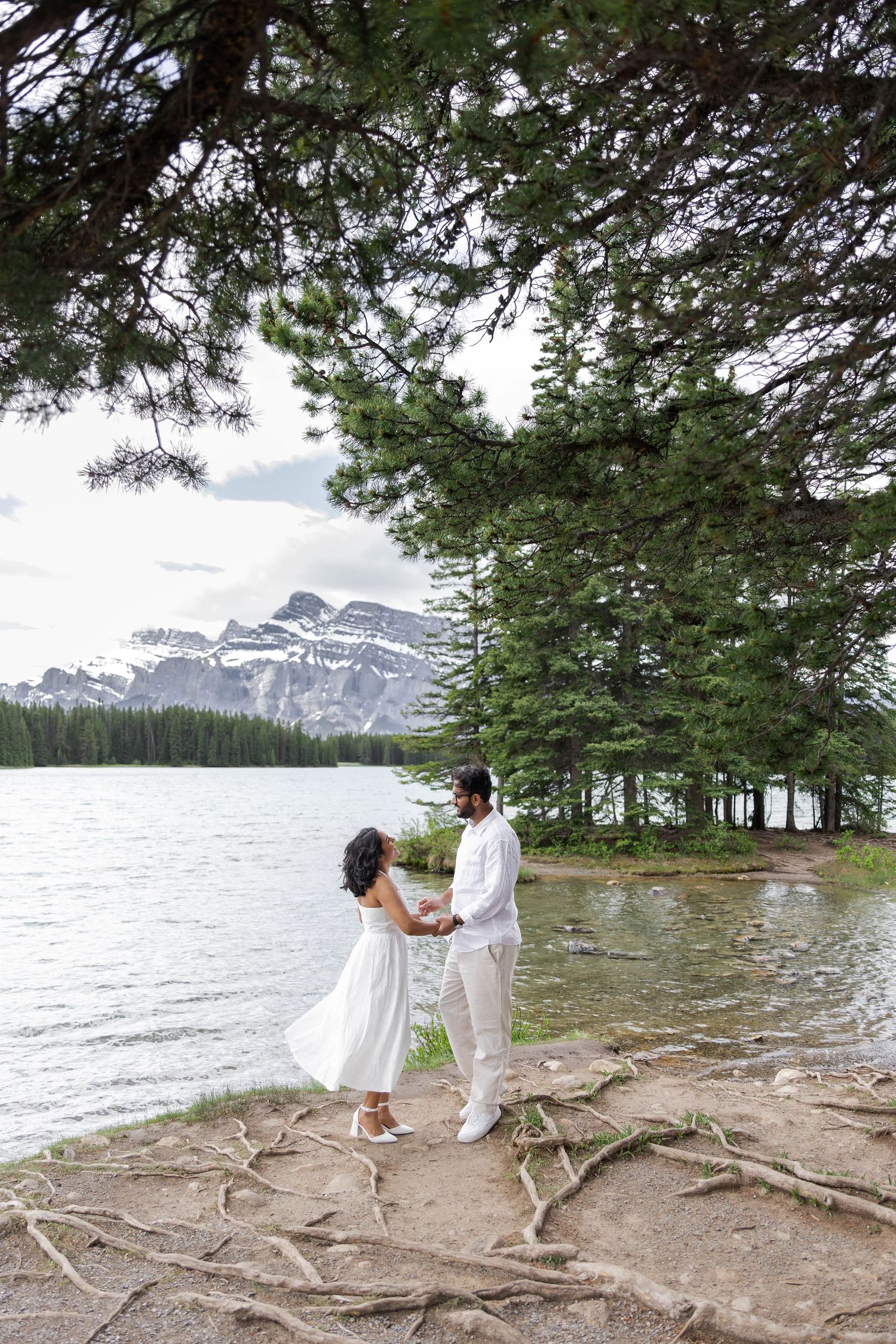 Couple in white holding hands by a mountain Two Jack Lake surrounded by pine trees and exposed roots.