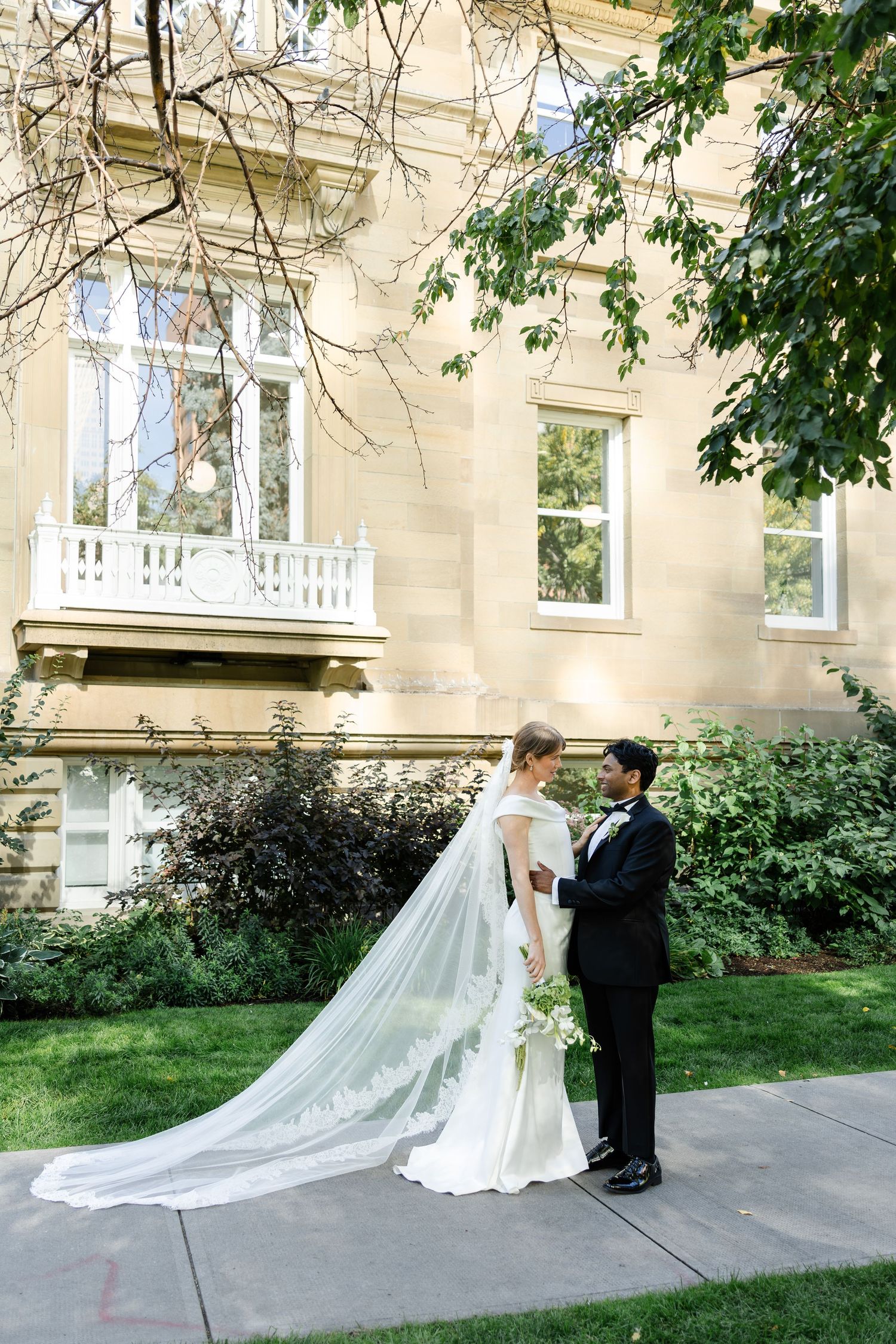 A bride and groom share a tender moment outside a grand historic building with lush gardens.