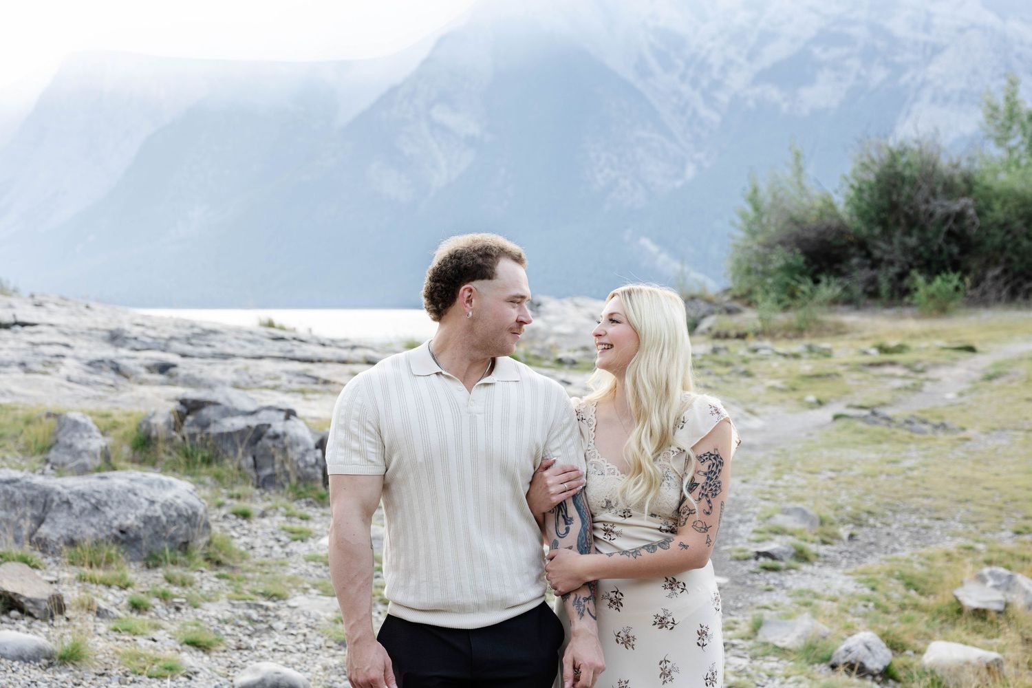 Happy couple walking arm in arm along a rocky mountain lakeside trail with misty peaks behind.Romantic couple in Minnewanka Lake in Banff.