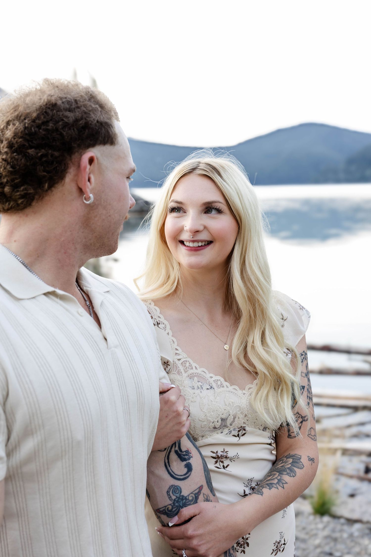 Tattooed blonde woman smiling at her partner near a serene mountain lake with rolling hills behind.Romantic couple in Minnewanka Lake