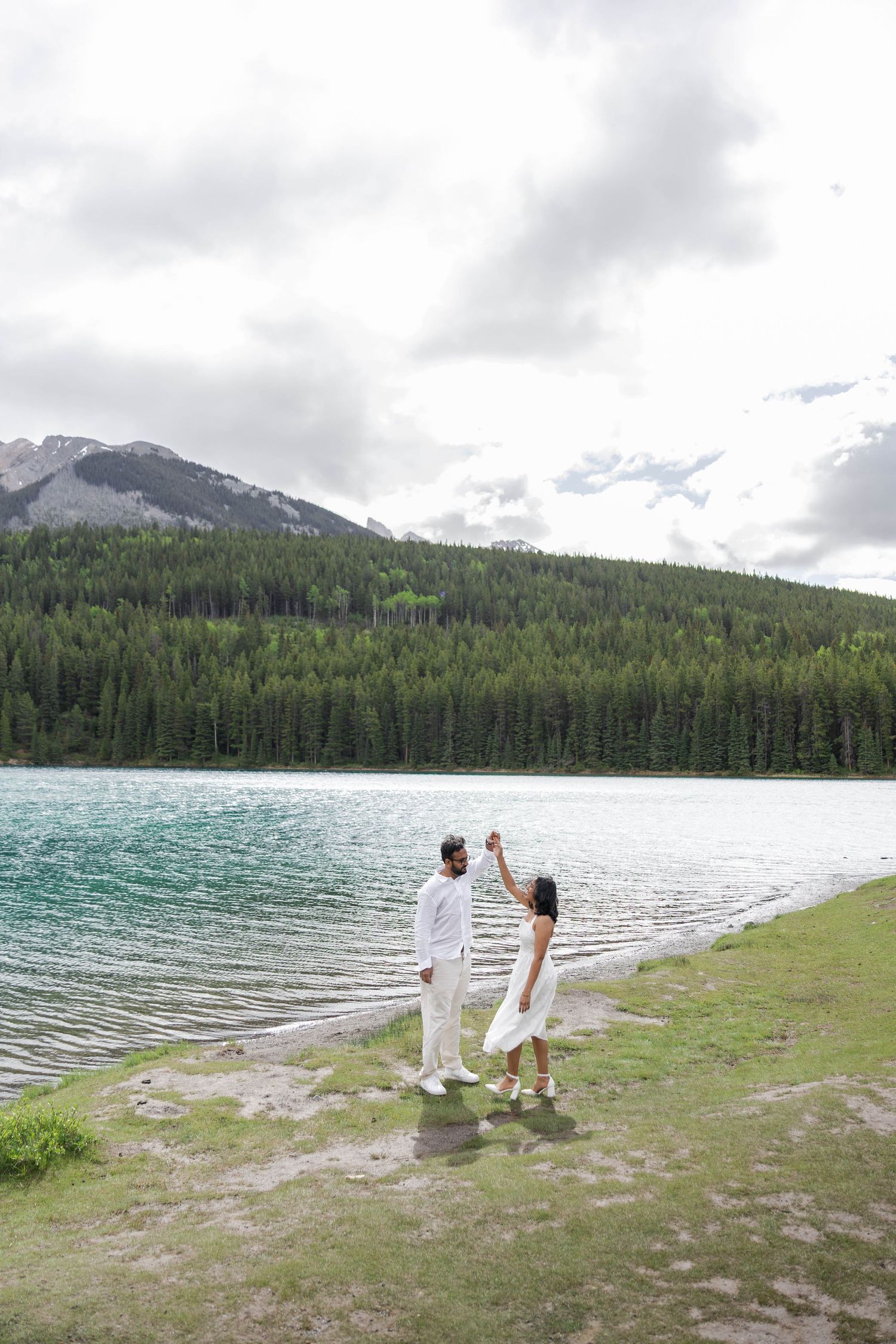 Couple dancing in white outfits by turquoise mountain lake with pine forest backdrop in Banff.