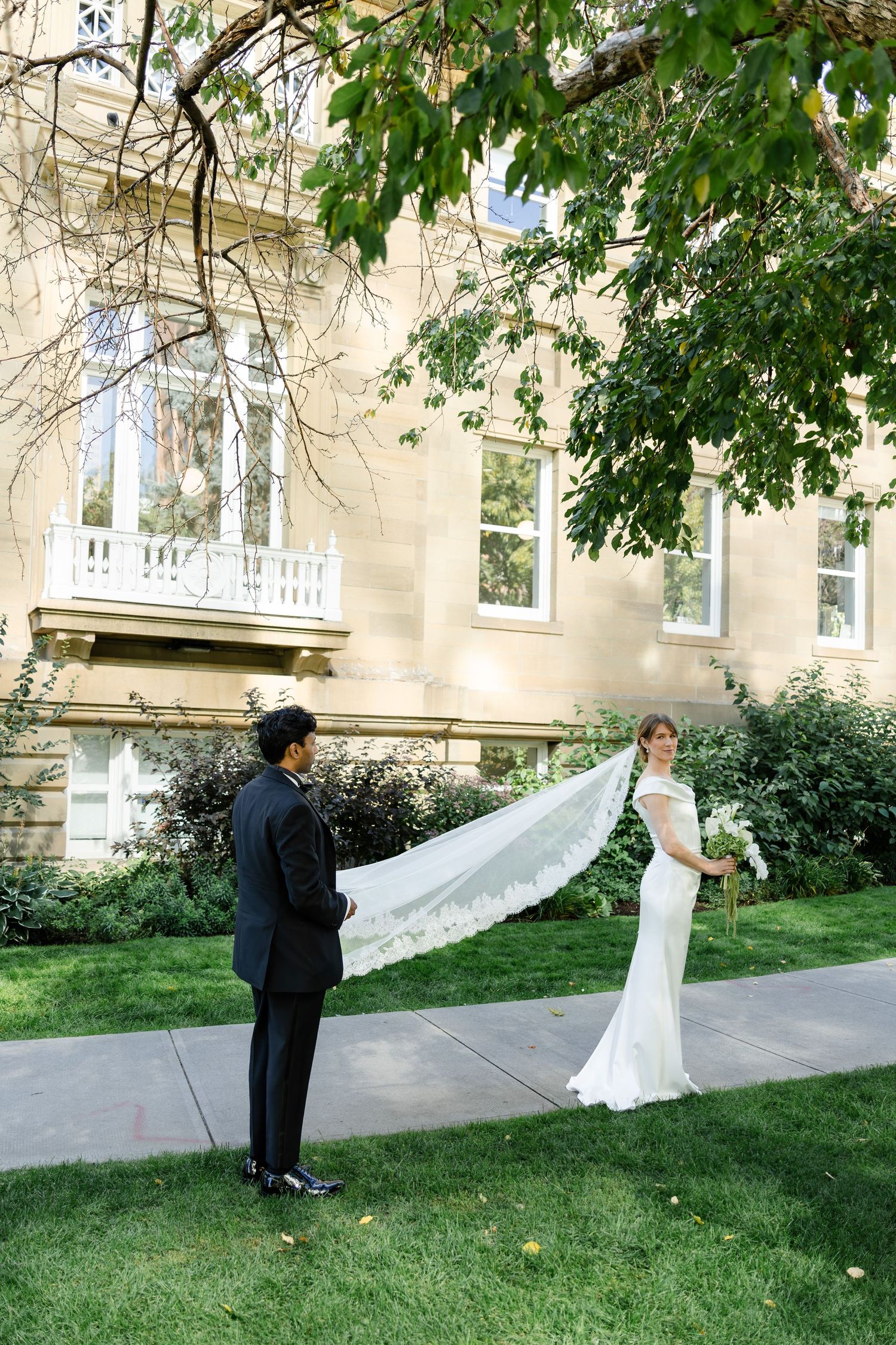 A bride in a flowing veil stands before a groom outside a historic stone building.