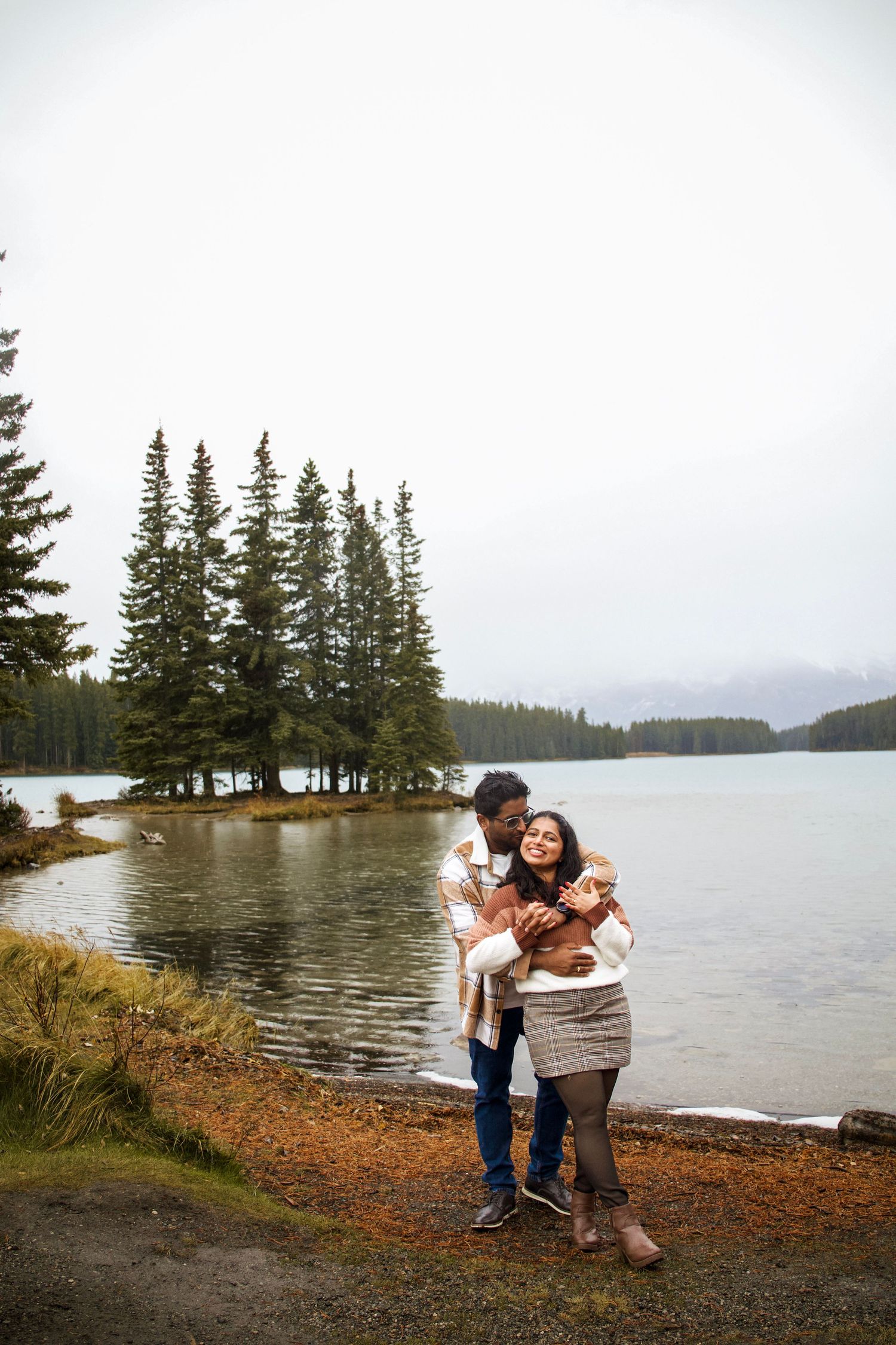 Couple embracing and kissing by a scenic mountain lake surrounded by evergreen trees on a cloudy day.