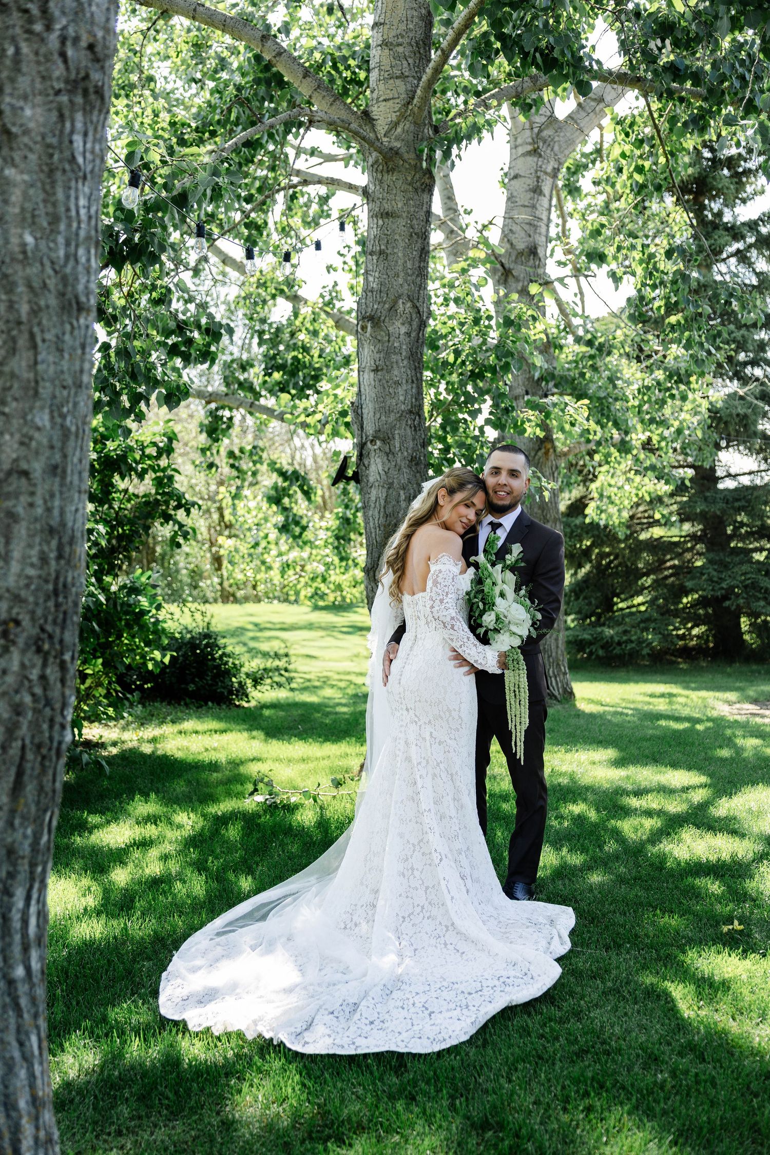 Bride in lace gown and groom in suit embrace under large trees on green lawn during outdoor wedding.