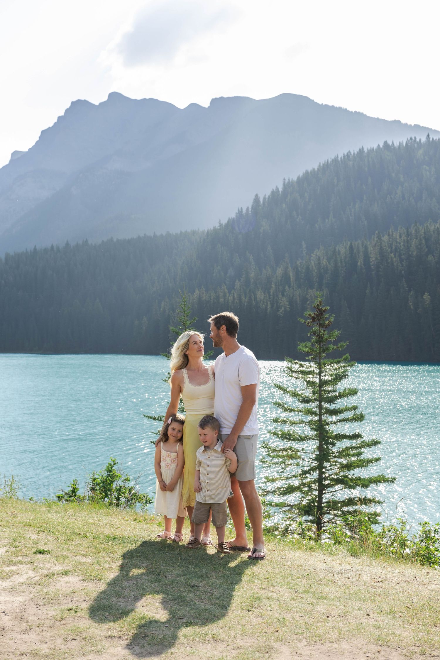 Family of four posing by a turquoise mountain Lake Minnewanka surrounded by pine trees and rocky peaks on a sunny day in Banff