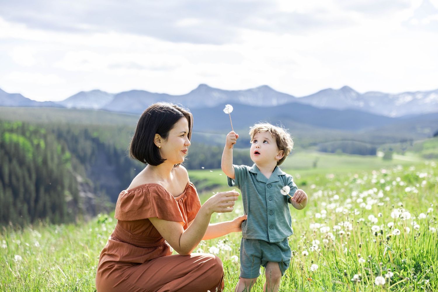 A mother and toddler boy blowing dandelion seeds in a green meadow with mountain views on a sunny day.
