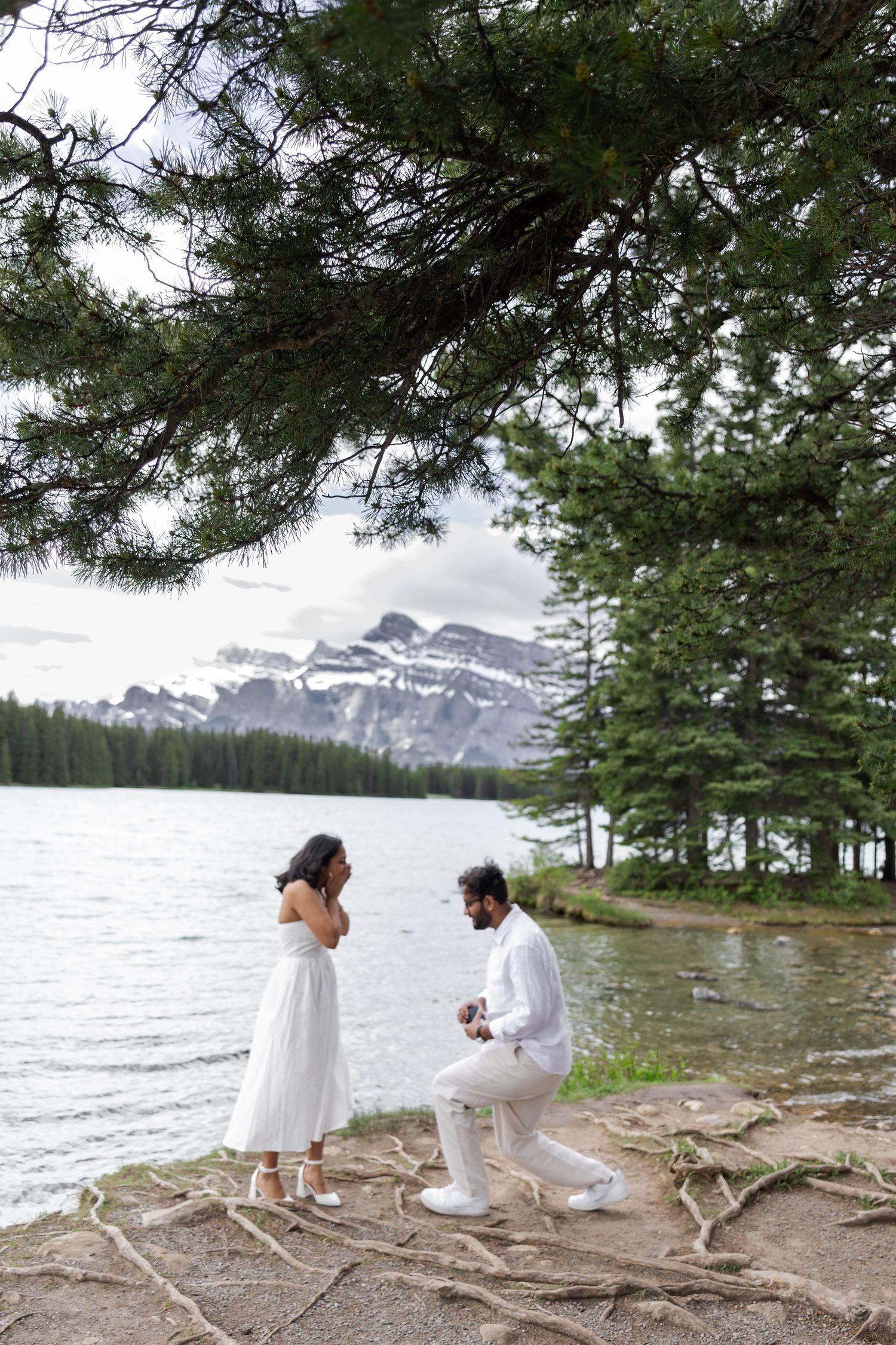 Man proposing to woman in white dress by a mountain lake, surrounded by pine trees and snow-capped peaks.