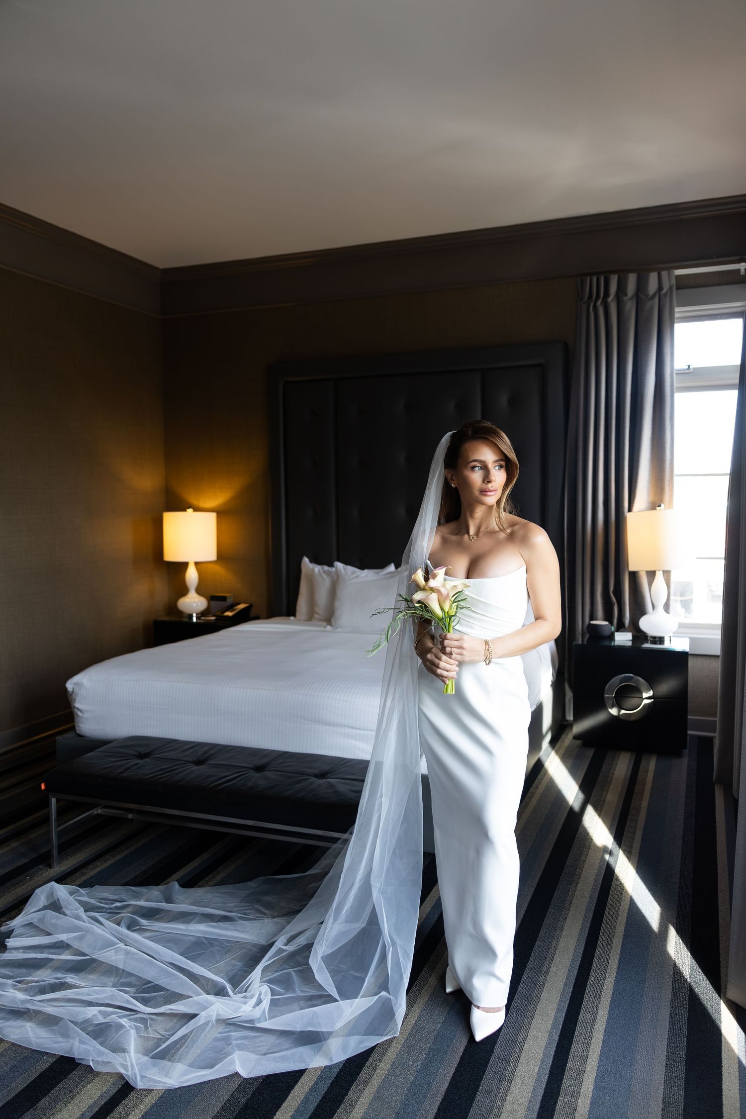 Elegant bride in white strapless gown and long veil holding floral bouquet standing in a luxurious hotel bedroom.