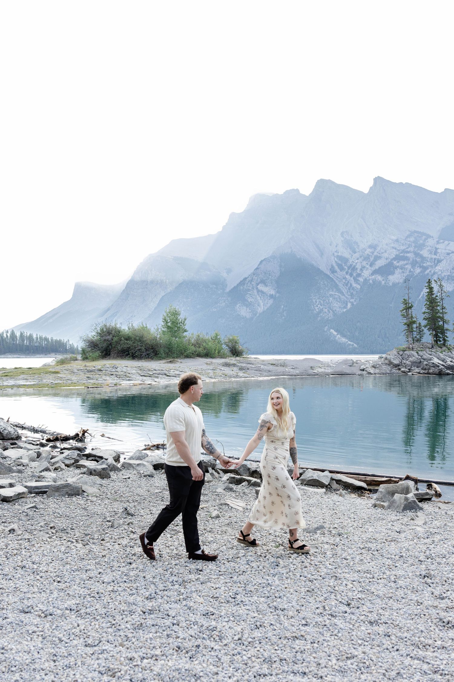 Couple holding hands walking along rocky lakeshore with turquoise water and snow-capped mountains in Banff.Romantic couple in Minnewanka Lak