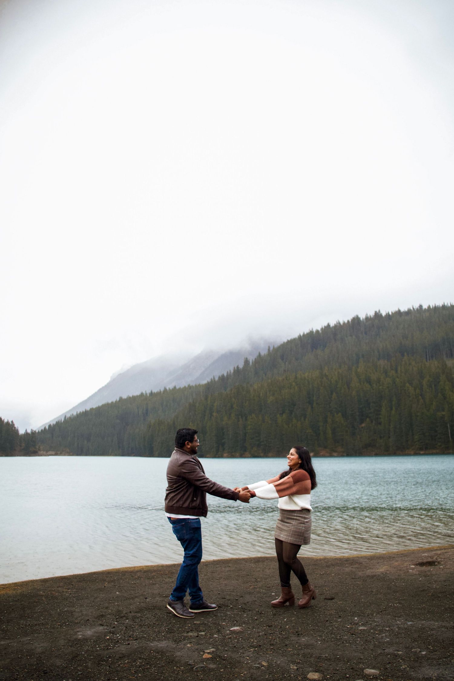Couple holding hands by a misty mountain lake surrounded by evergreen trees on an overcast day.
