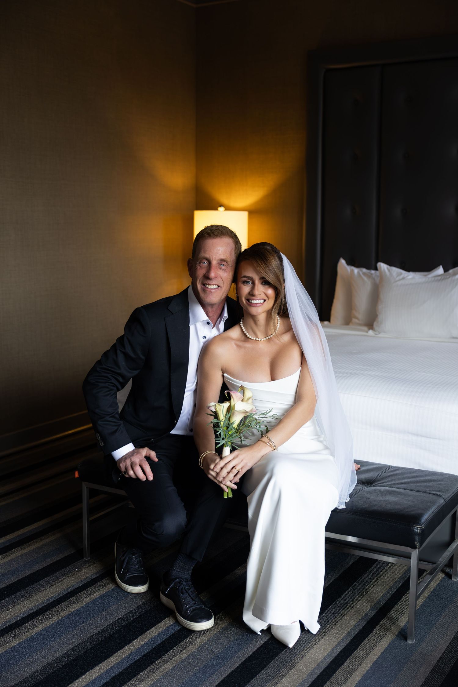 A smiling bride in white gown with veil holding bouquet sits beside groom in black suit on hotel room bench.