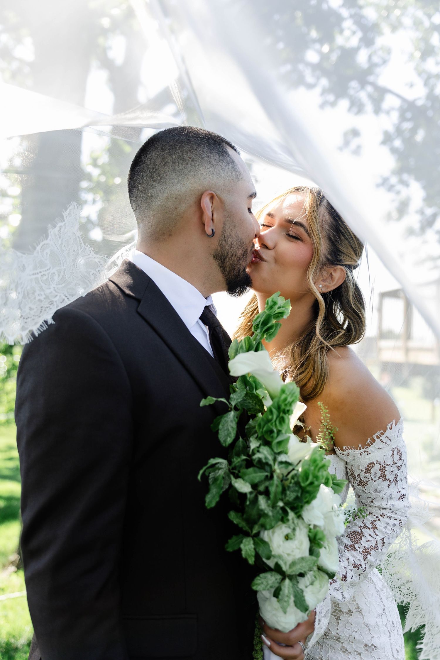 Bride and groom sharing a romantic kiss under a white lace veil, bride holding a white and green bouquet.
