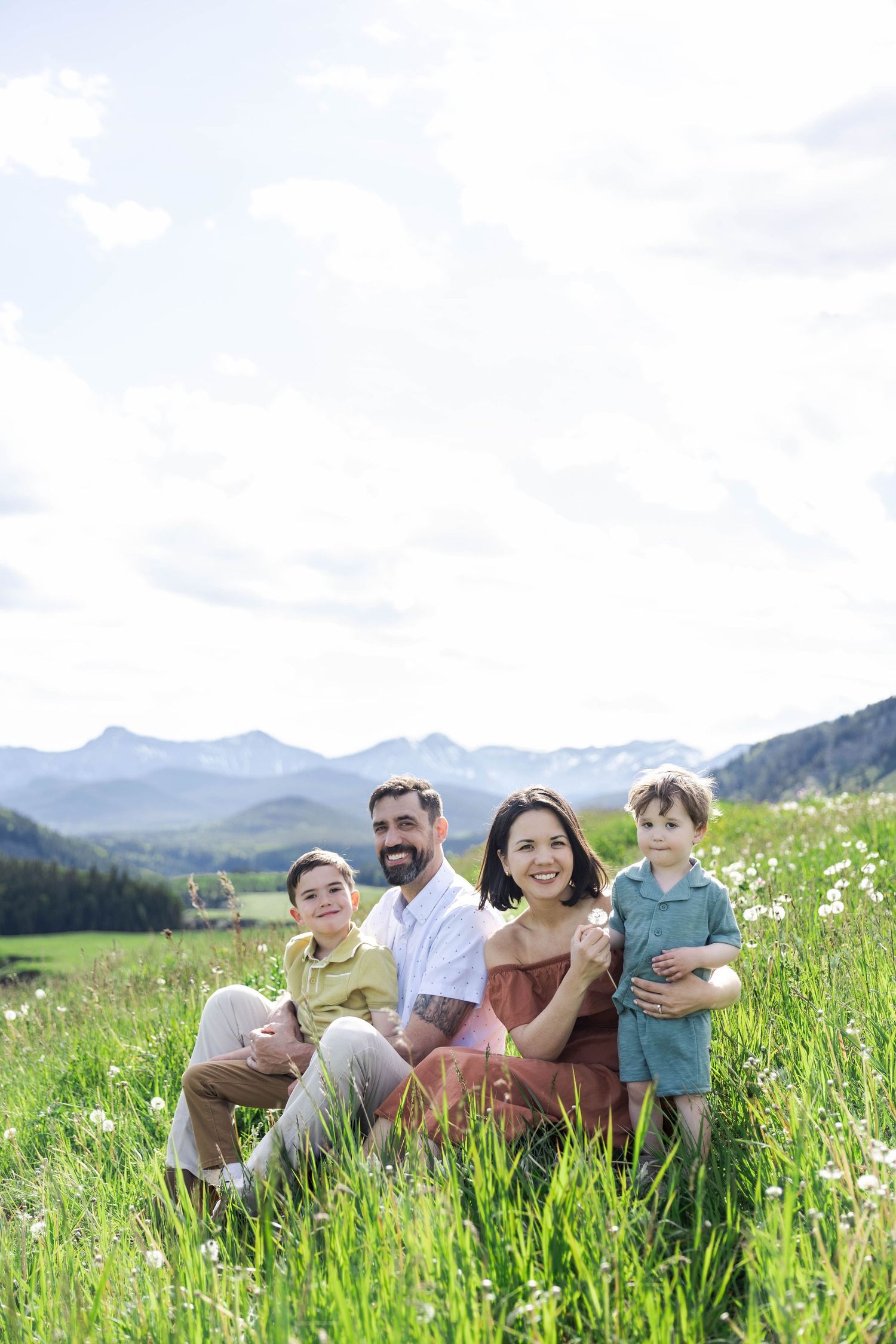 Family of four sitting in green meadow with Kananaskis mountain backdrop on sunny day, smiling at camera.