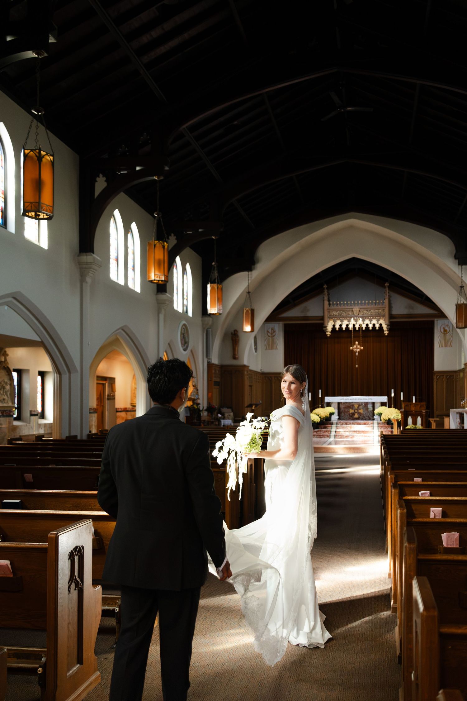 Bride in white gown holds hands with groom in black suit inside a sunlit church aisle.