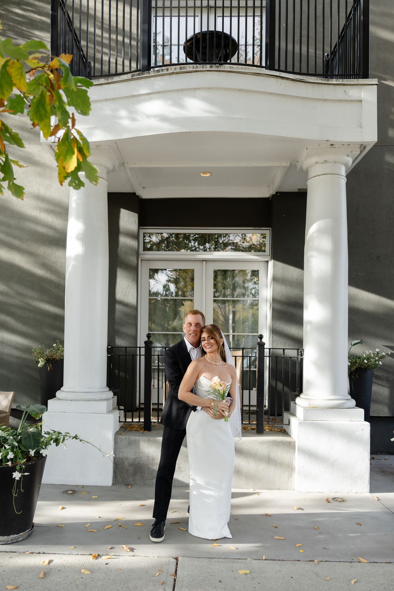 A smiling bride and groom pose in front of an elegant building with white columns on their wedding day.