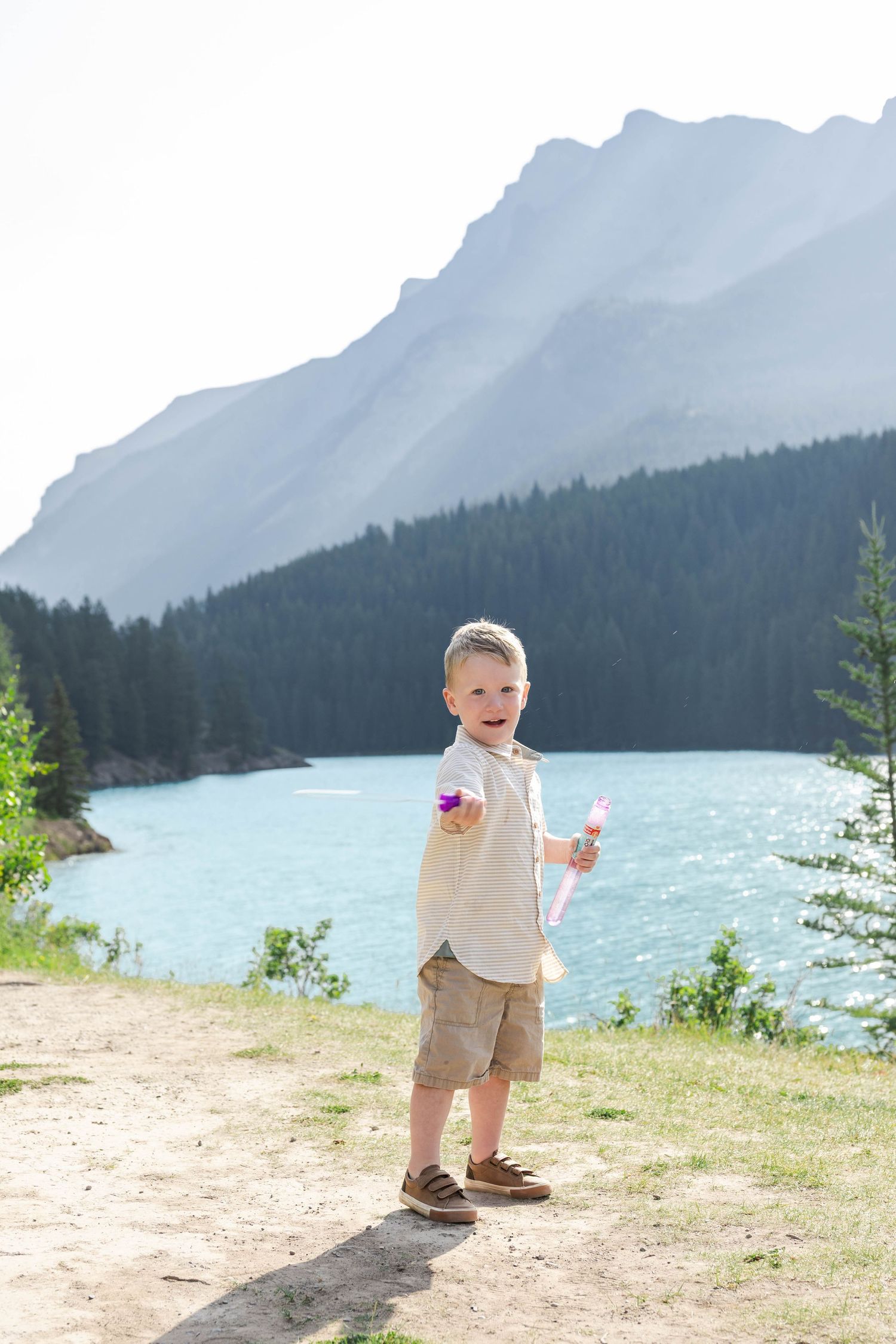 Young boy holding bubble wand by turquoise mountain Lake Minnewanka with pine forest and rocky peaks in background on sunny day.in Banff