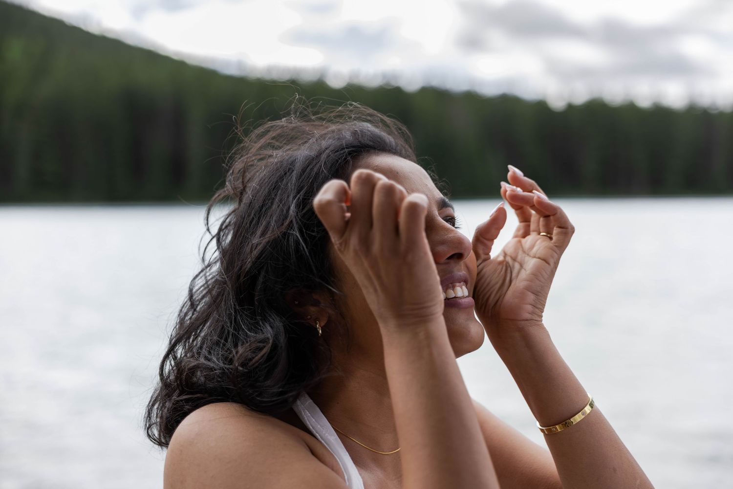 Smiling woman holding binoculars near a serene lake surrounded by lush green forest on a cloudy day.