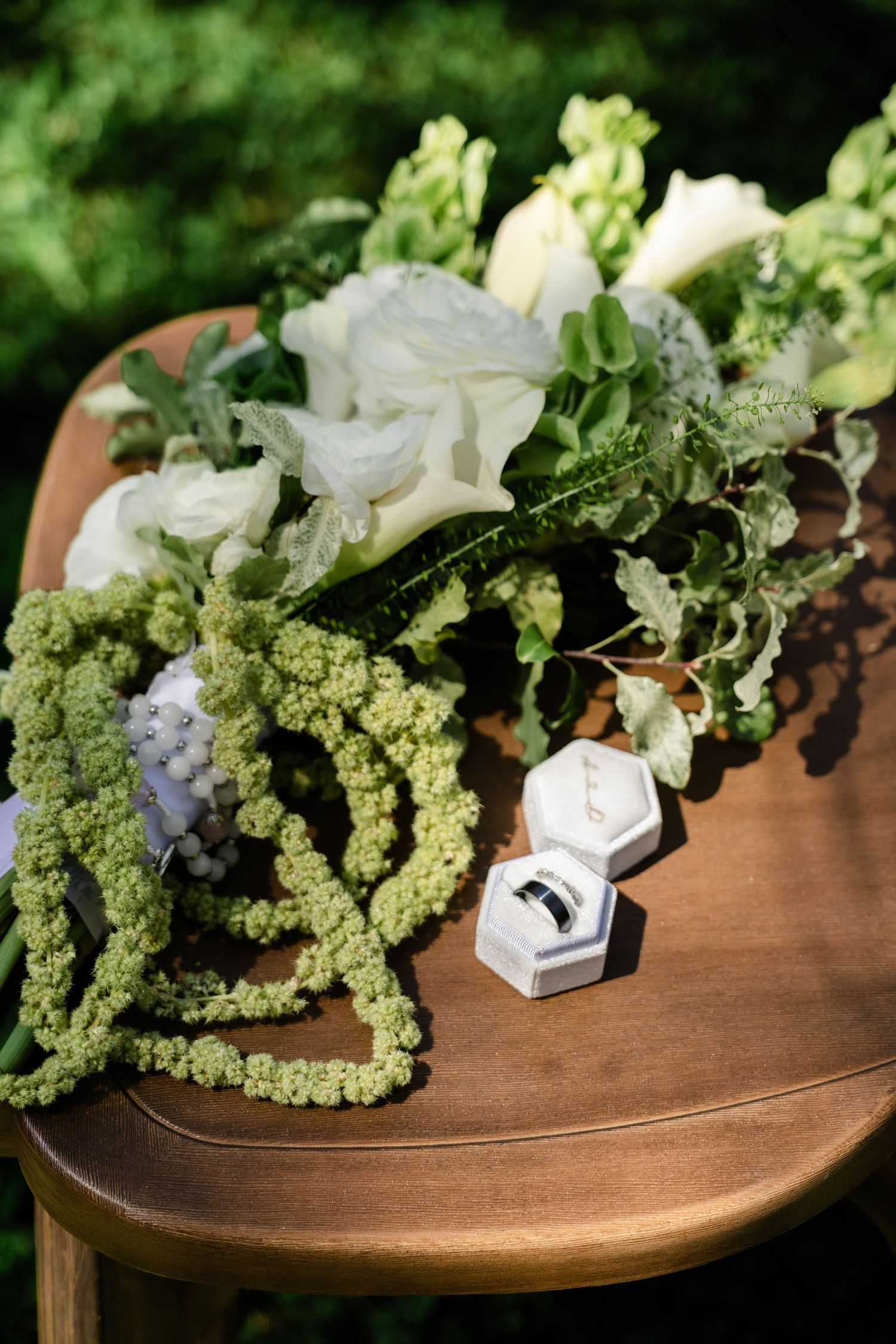 Wedding bouquet with white flowers and green accents beside a ring box on a wooden chair.