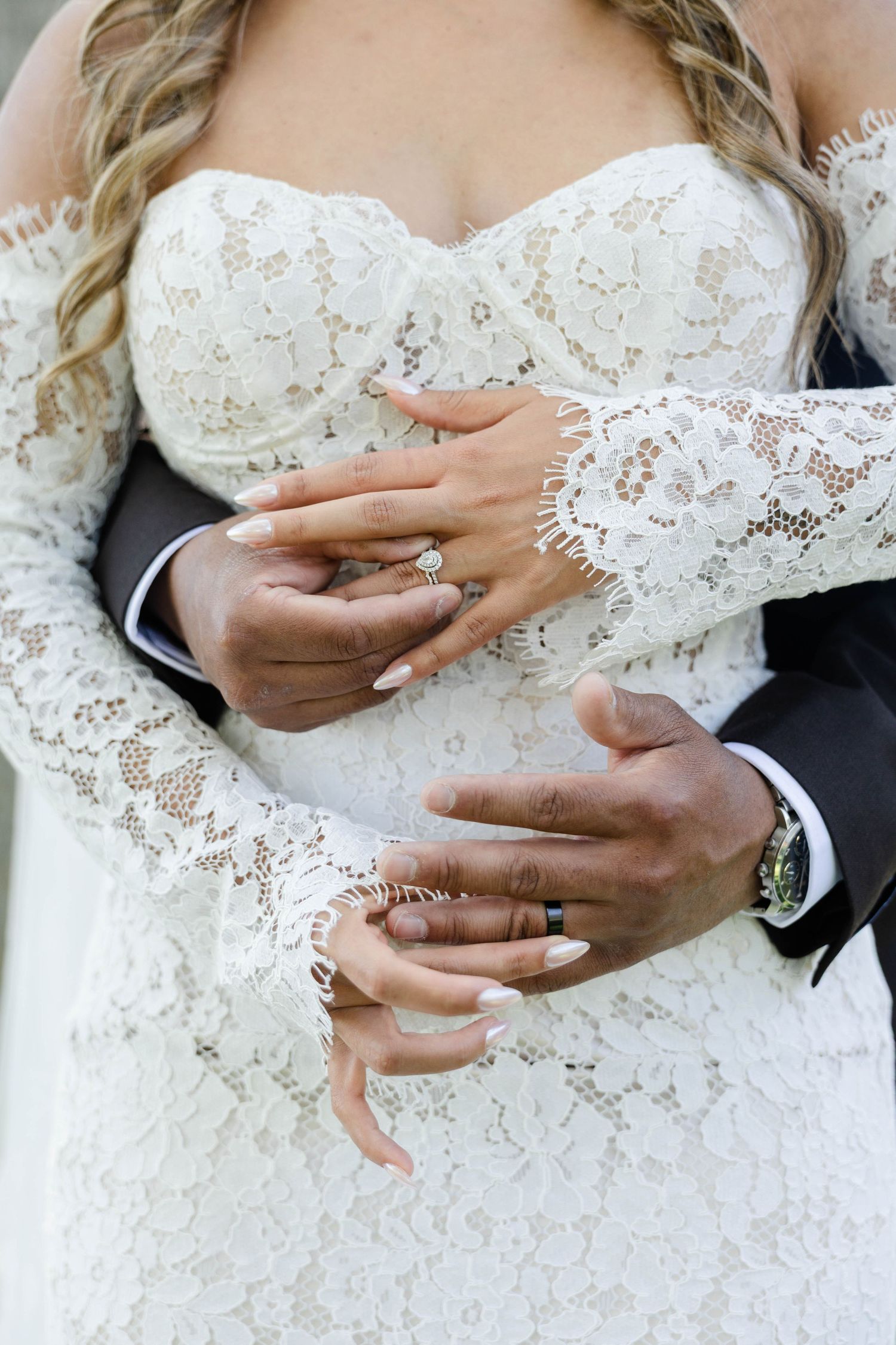 Interracial newlyweds holding hands showing wedding rings, bride in white lace gown embraced by groom in black suit.