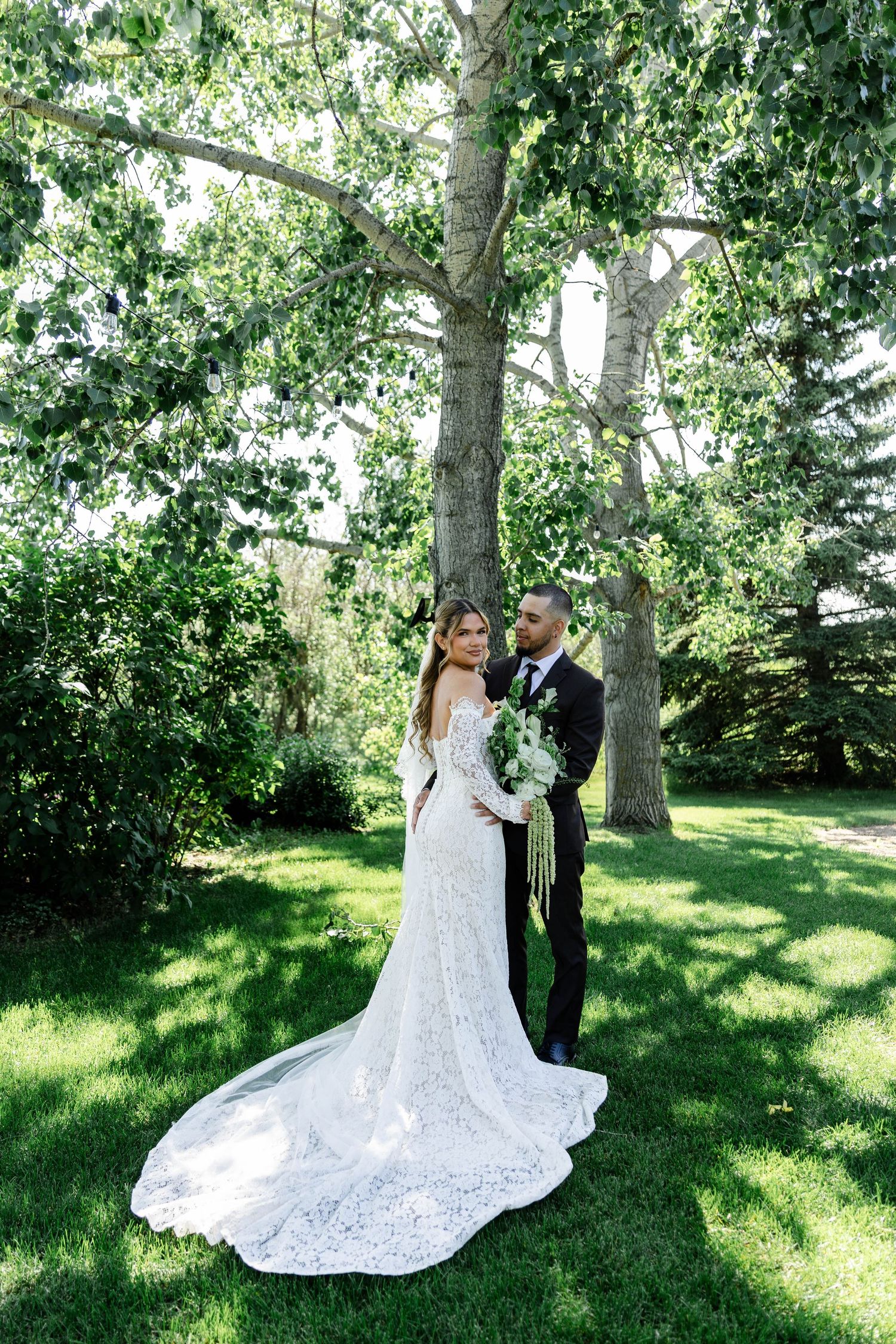 Bride in lace gown with train and groom in dark suit pose together on green lawn beneath large trees.