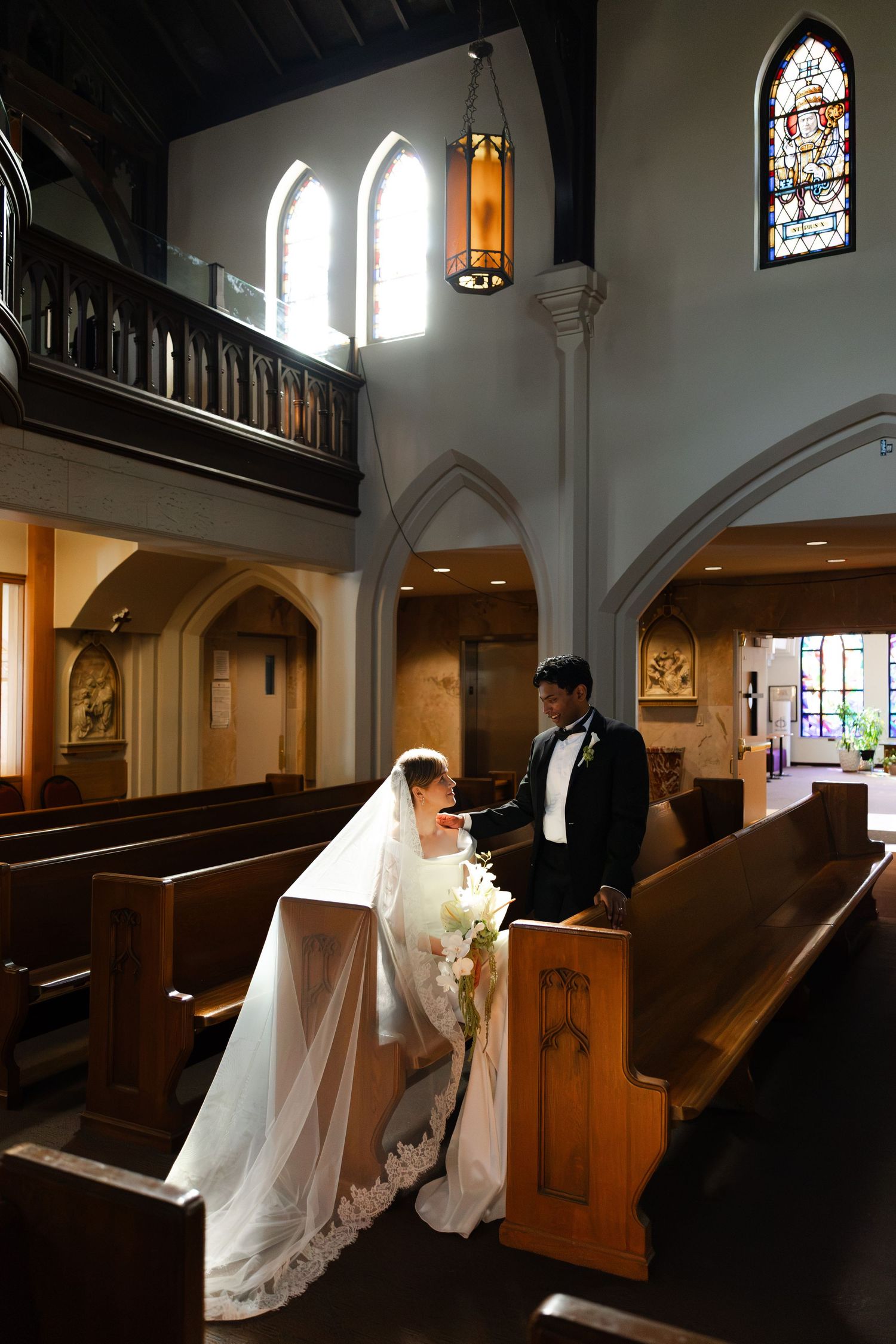 Bride and groom share a tender moment in a church aisle, with stained glass windows and wooden pews.