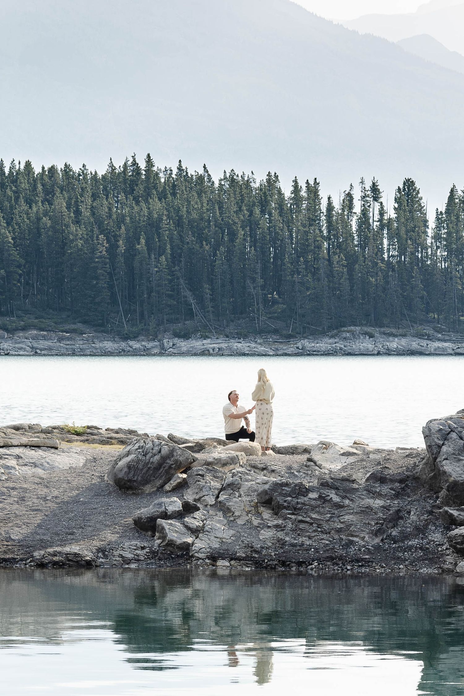 Man proposing on rocky lakeside with evergreen forest and mountains reflected in calm water.Romantic couple in Minnewanka Lake in Banff.