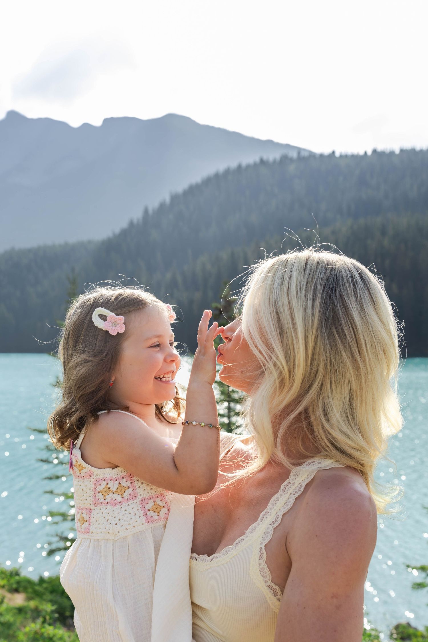 A blonde woman holds a smiling little girl with a pink flower clip near a turquoise mountain Lake Minnewanka in Banff