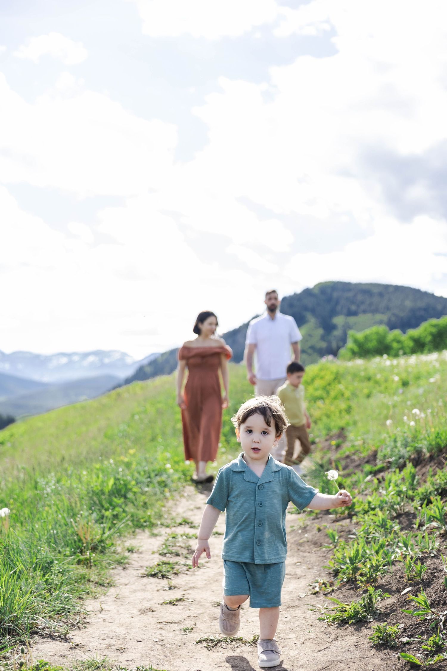 Young boy walks down a mountain trail as his family follows on a sunny green hillside.