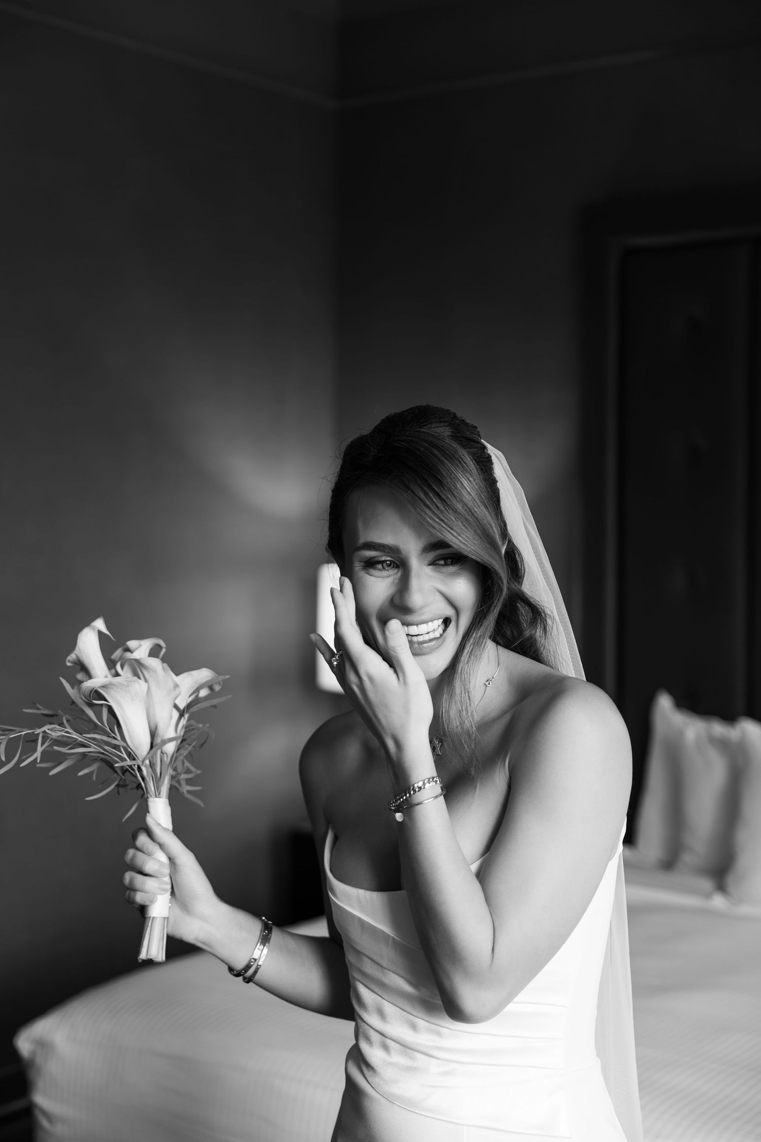 Black and white photo of a joyful bride in a white gown holding a bouquet, laughing and touching her face.