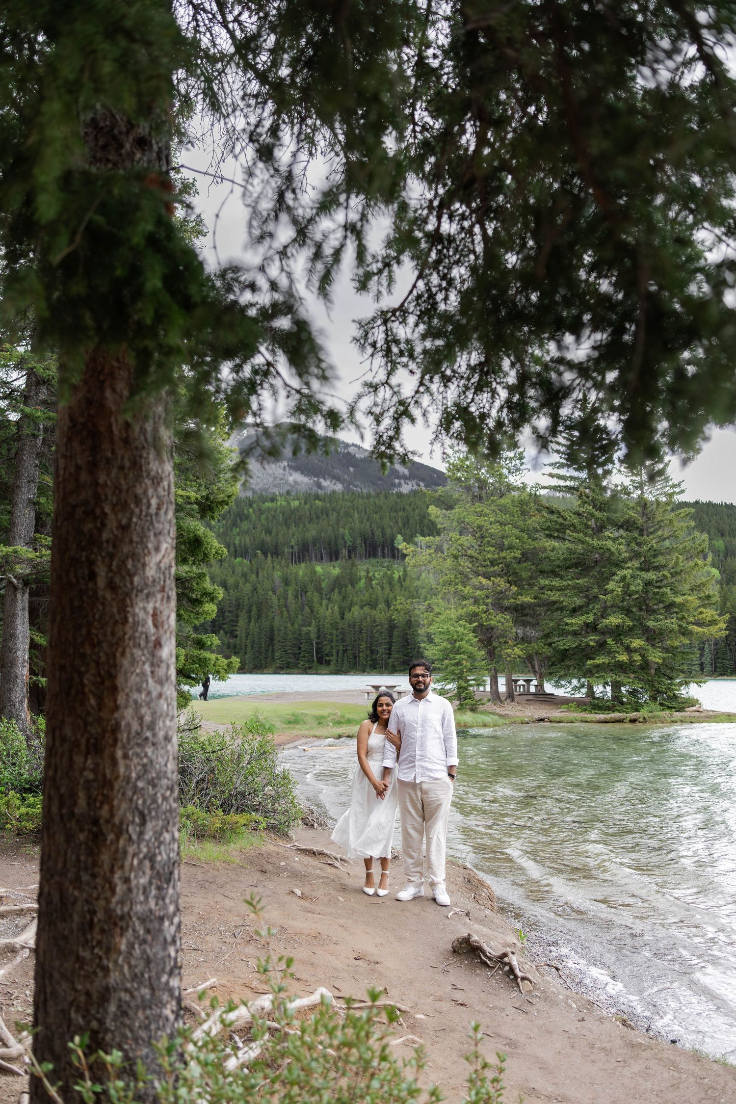 Couple in white wedding attire standing by a lakeside forest path with mountain views in the background.